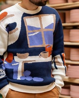 Alex wears a sweater with an illustration of a pottery workshop with a large window, shelves, and table with mugs and bowls. Alex stands in front of a rack of ceramic bowls in progress.