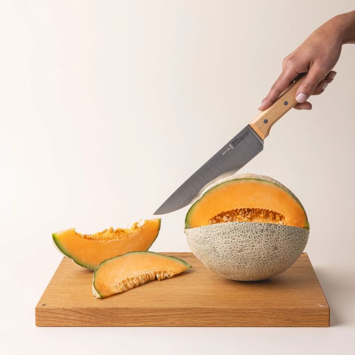 A hand holds a chef's knife with a steel blade and a light wooden handle and is cutting open a cantaloupe on a wooden cutting board.