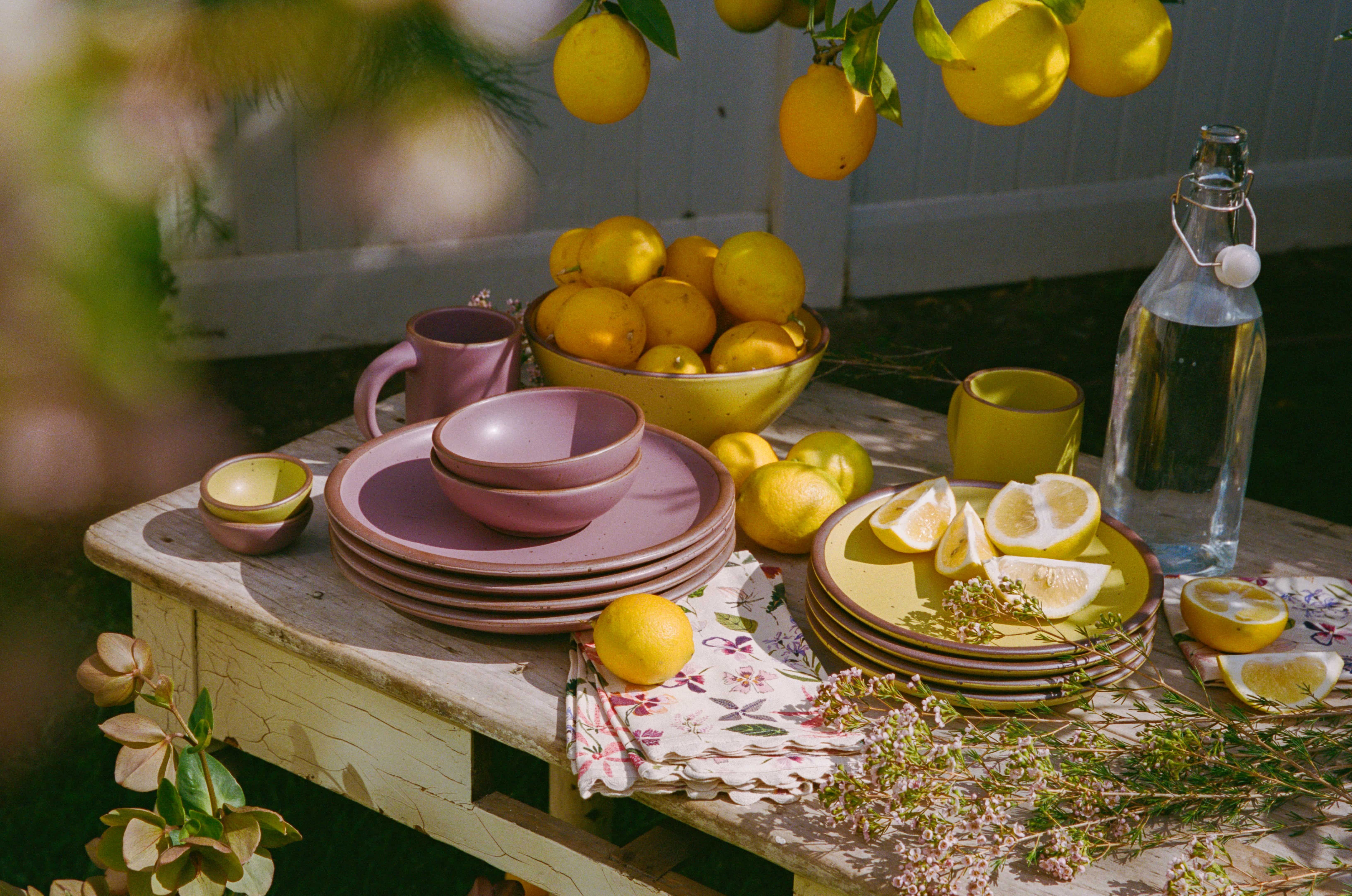 An outside table filled with ceramic dinnerware in zesty yellow and soft mauve purple colors, lemons, floral napkins, and flowers.