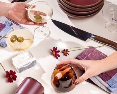 Hands reach for a martini coupe and a whiskey snifter against a white background. All around are other items including a stack of shallow bowls, kitchen knives, a plum ceramic mug, and more.