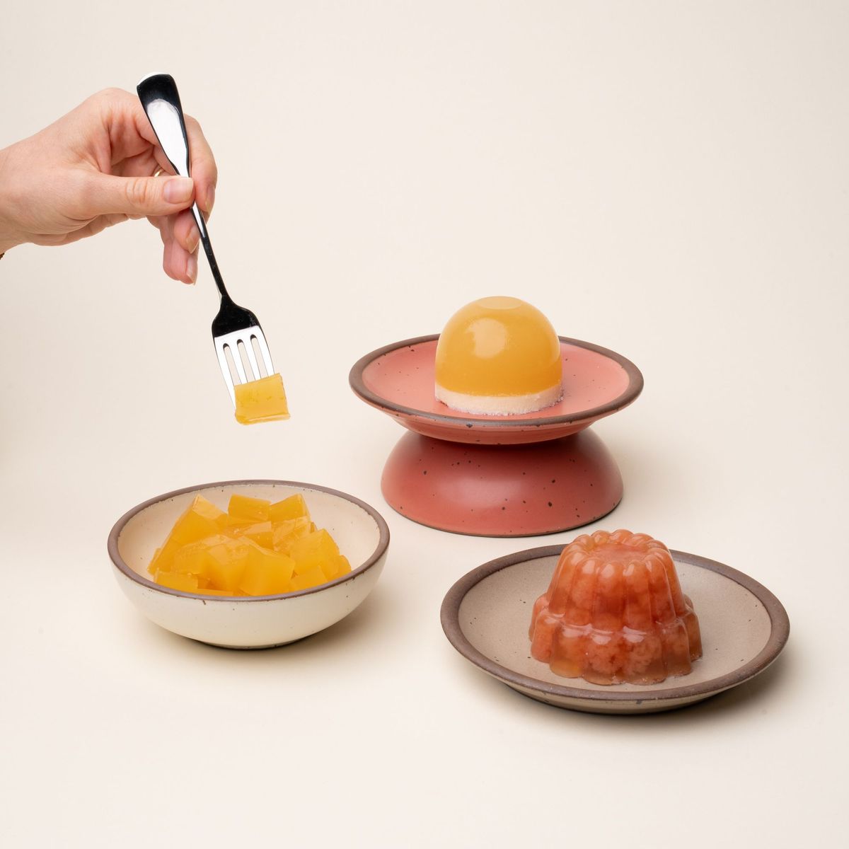Three colorful gelatin desserts on ceramic dishes: yellow cubes, a dome-shaped yellow and white layered gelatin on a pink stand, and a molded red gelatin, a hand holds a fork with a yellow cube of gelatin.