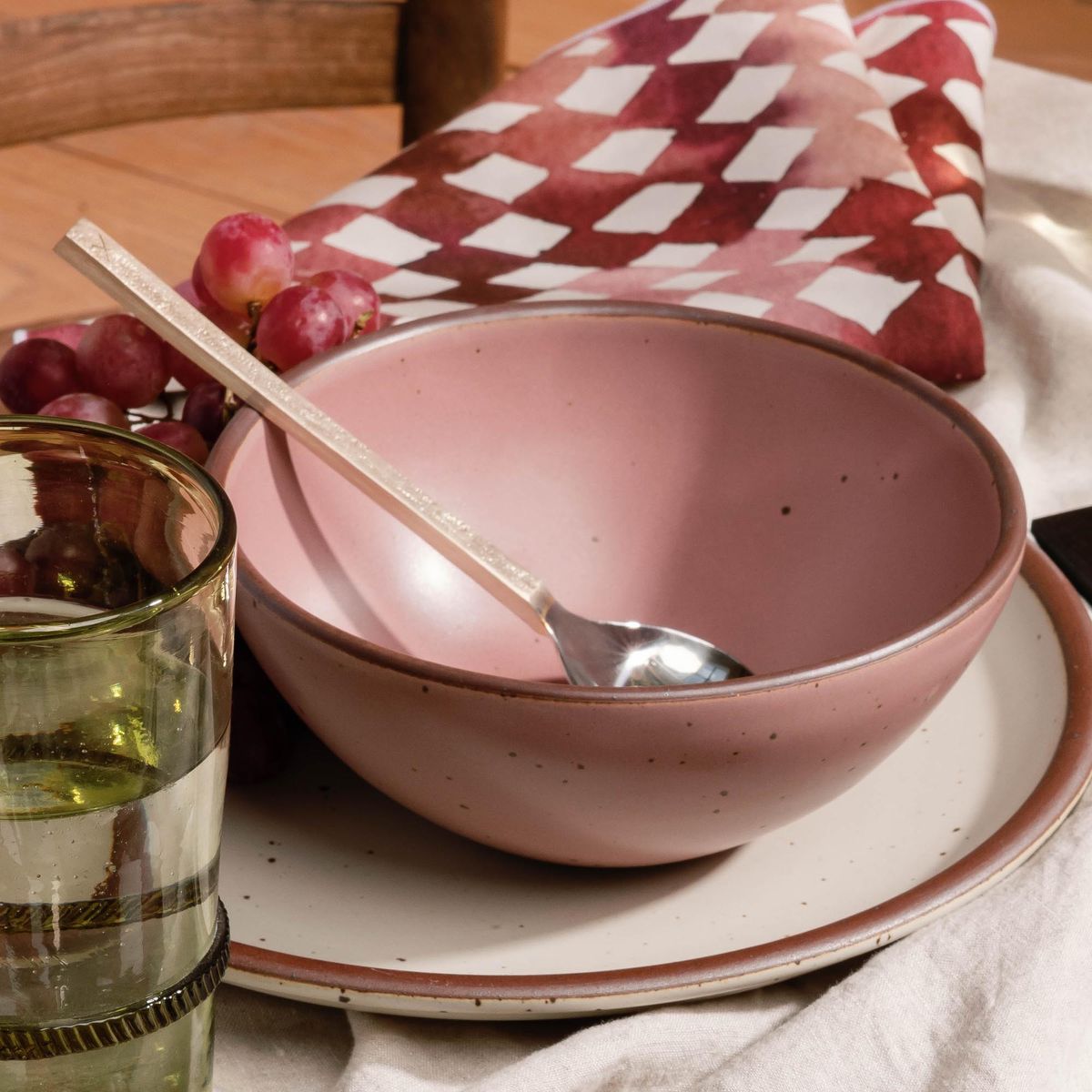 Dusty mauve pink ceramic bowl with a spoon resting inside, set on a plate with grapes and a patterned napkin nearby.