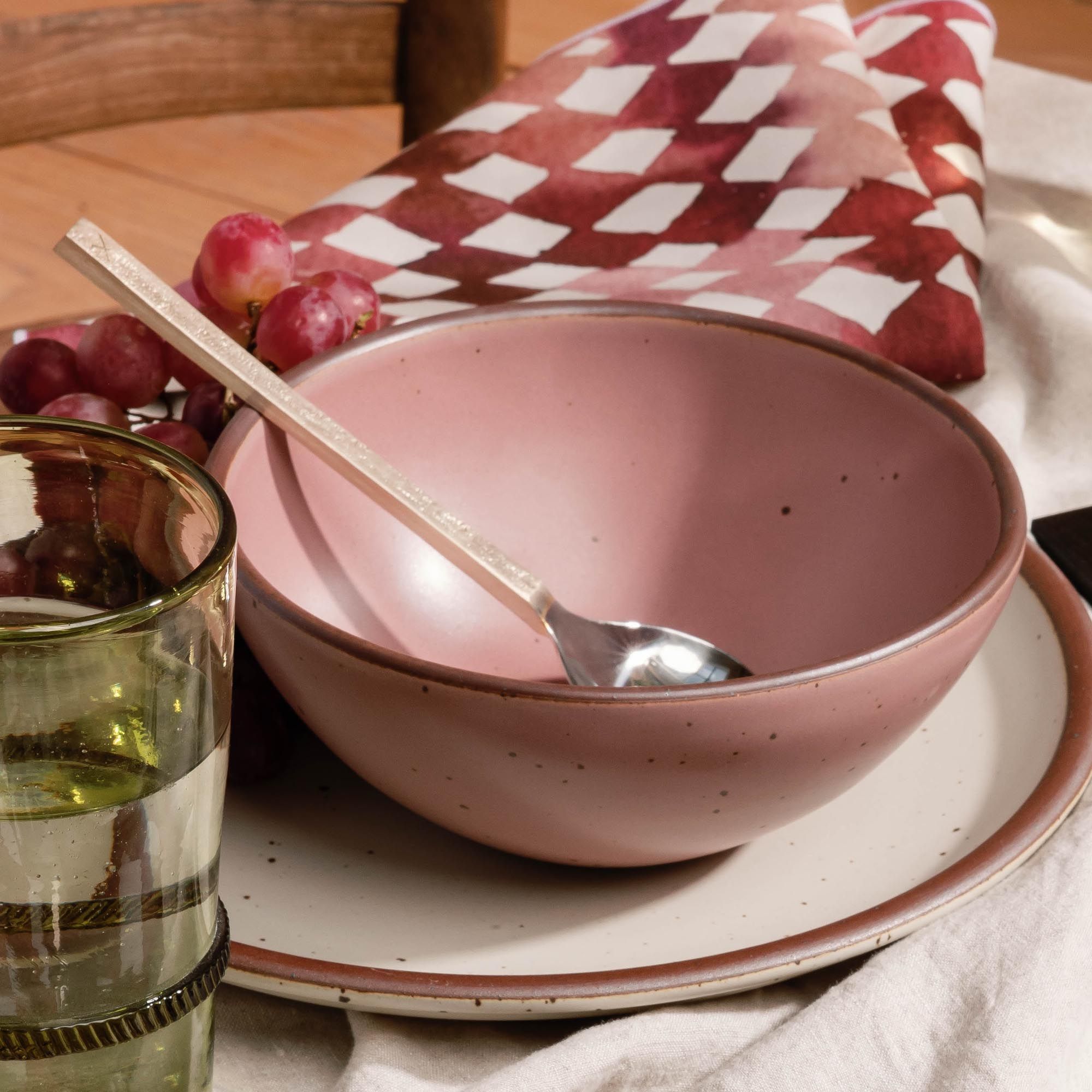Dusty mauve pink ceramic bowl with a spoon resting inside, set on a plate with grapes and a patterned napkin nearby.