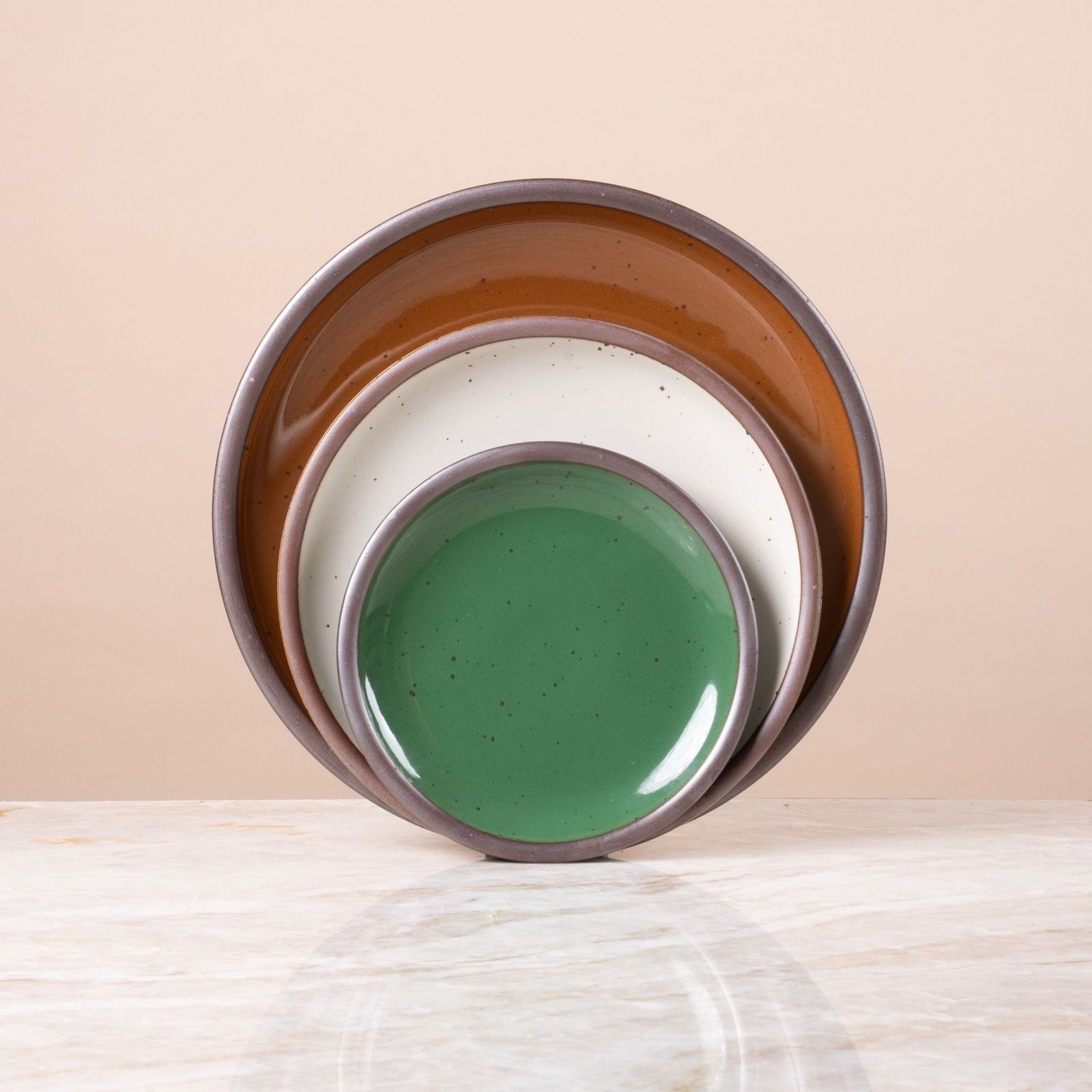 Stacked ceramic plates in glossy green, white, and glossy brown with speckled finish, displayed on a marble surface.