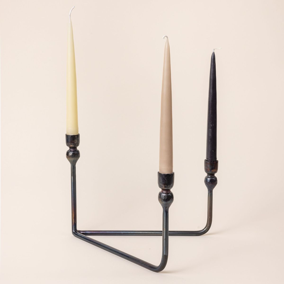 3 taper candles in a natural, taupe, and black colors sit in a sophisticated iron candelabra. The candelabra has a v shaped base and 3 candlestick holders.