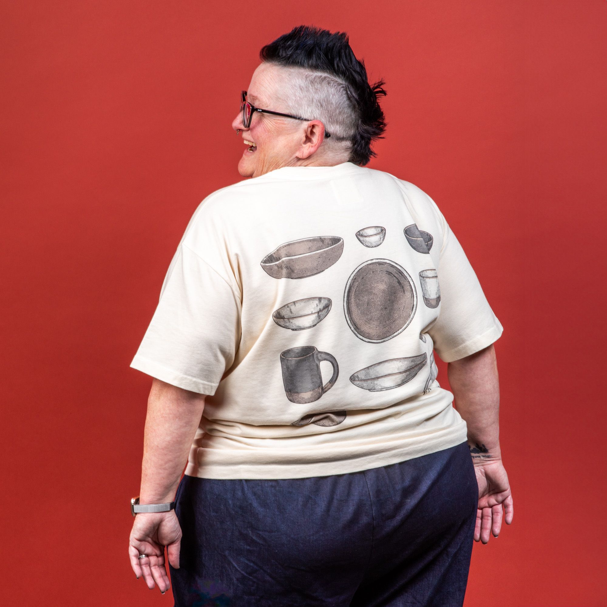 Model wearing the t-shirt from the back and staring off to the side smiling and wearing pink glasses