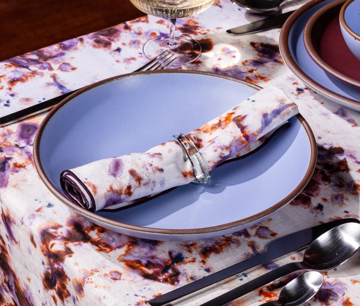 A rolled linen napkin in a marbled pattern with white, pink, purple, and orange colors sits on a ceramic plum dish.