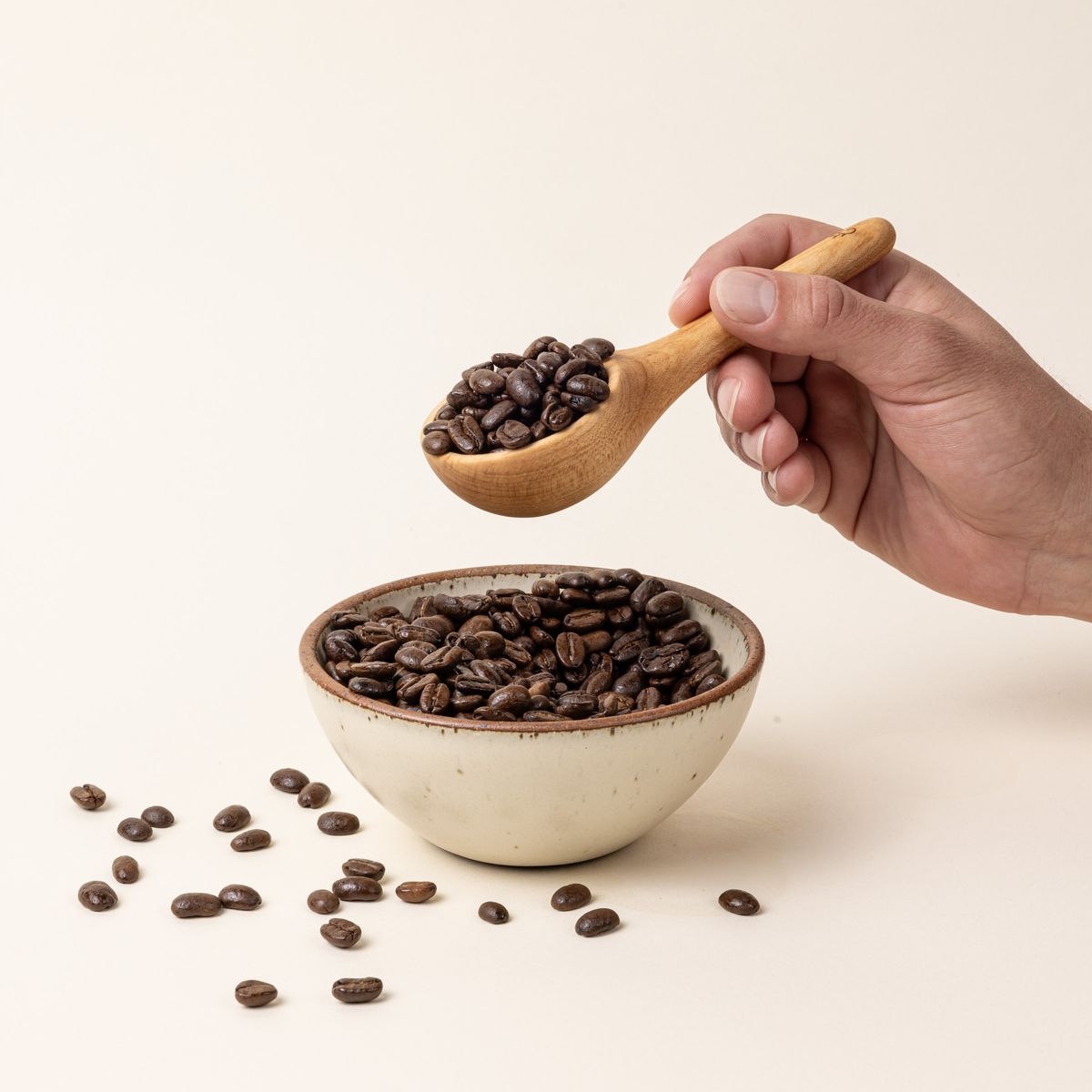 A hand holds a short wooden coffee scoop filled with coffee beans over a small bowl