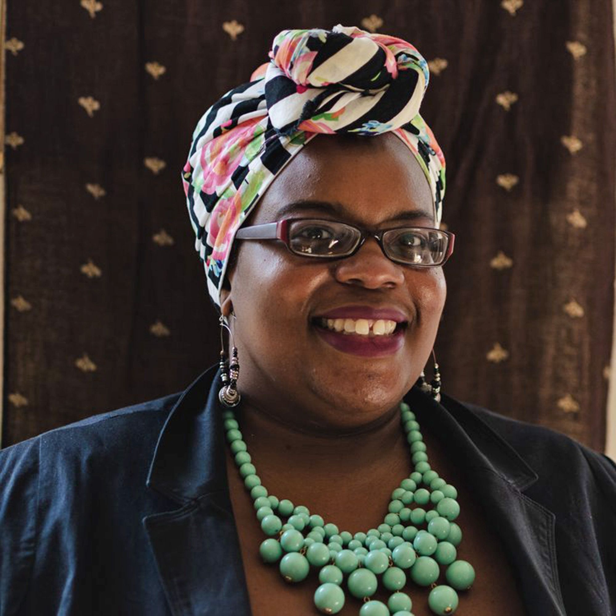 A person is smiling wearing a colorful headscarf and glasses and smiling at the camera