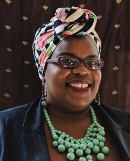 A person is smiling wearing a colorful headscarf and glasses and smiling at the camera