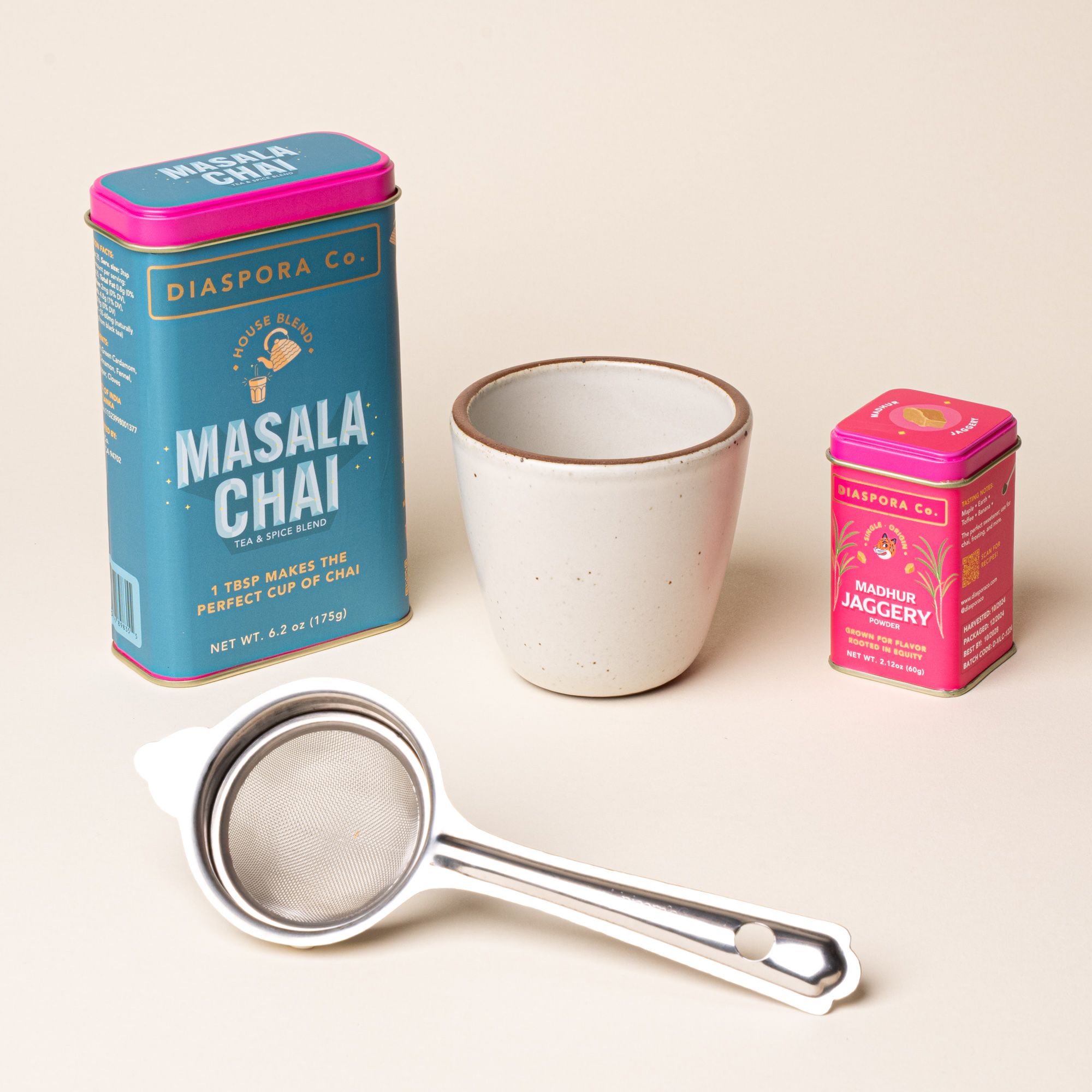 Masala chai tea kit with a ceramic cup in a warm off-white color, stainless steel strainer, and tins of Diaspora Co. masala chai blend and jaggery.