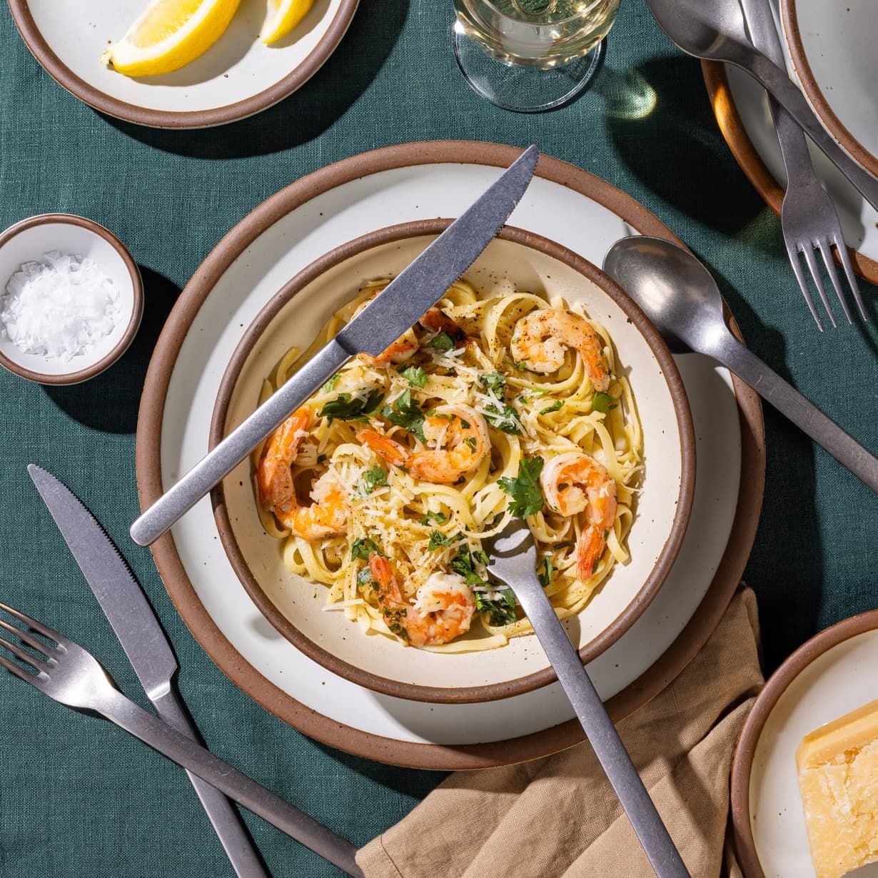 An off-white ceramic shallow bowl filled with pasta and shrimp, with steel flatware, and other ceramic cool white plates surrounding.