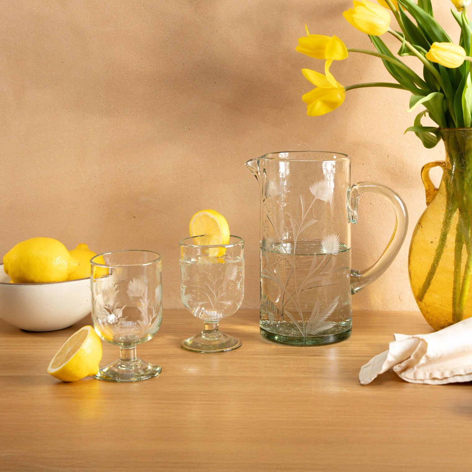 Etched thistle wine glasses and pitcher sit on a wooden table next to a bowl of lemons and vase of tulips.