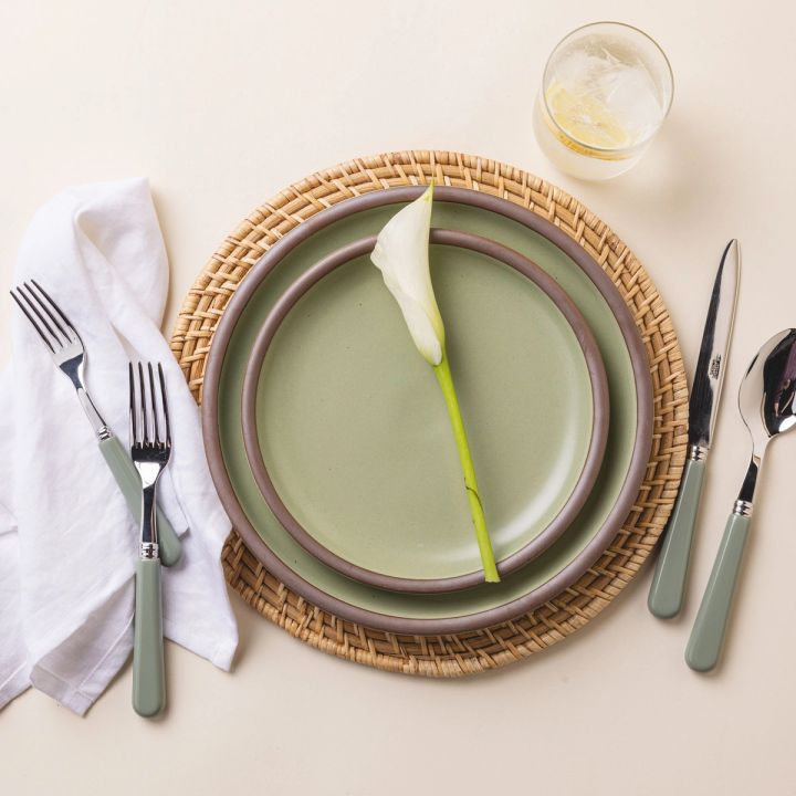 A place setting with 2 ceramics plates, 1 sage green, and 1 neutral, sitting on a woven natural charger. A sage matte handled fork and knife are arranged on the sides of the plates, along with a calla lily stem and a clear glass.