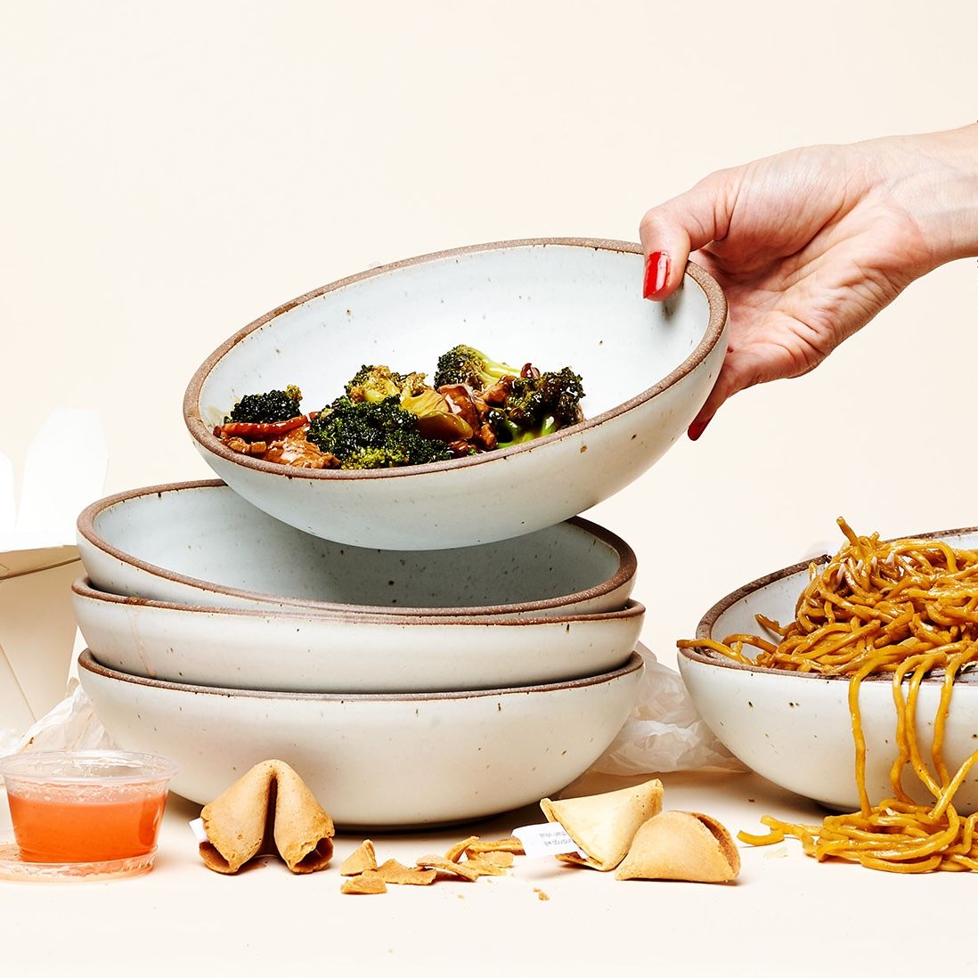 A hand holds a shallow bowl filled with cooked broccoli and about to stack on a pile of other shallow bowls. Surrounding is noodles, fortune cookies, and a little sauce container.