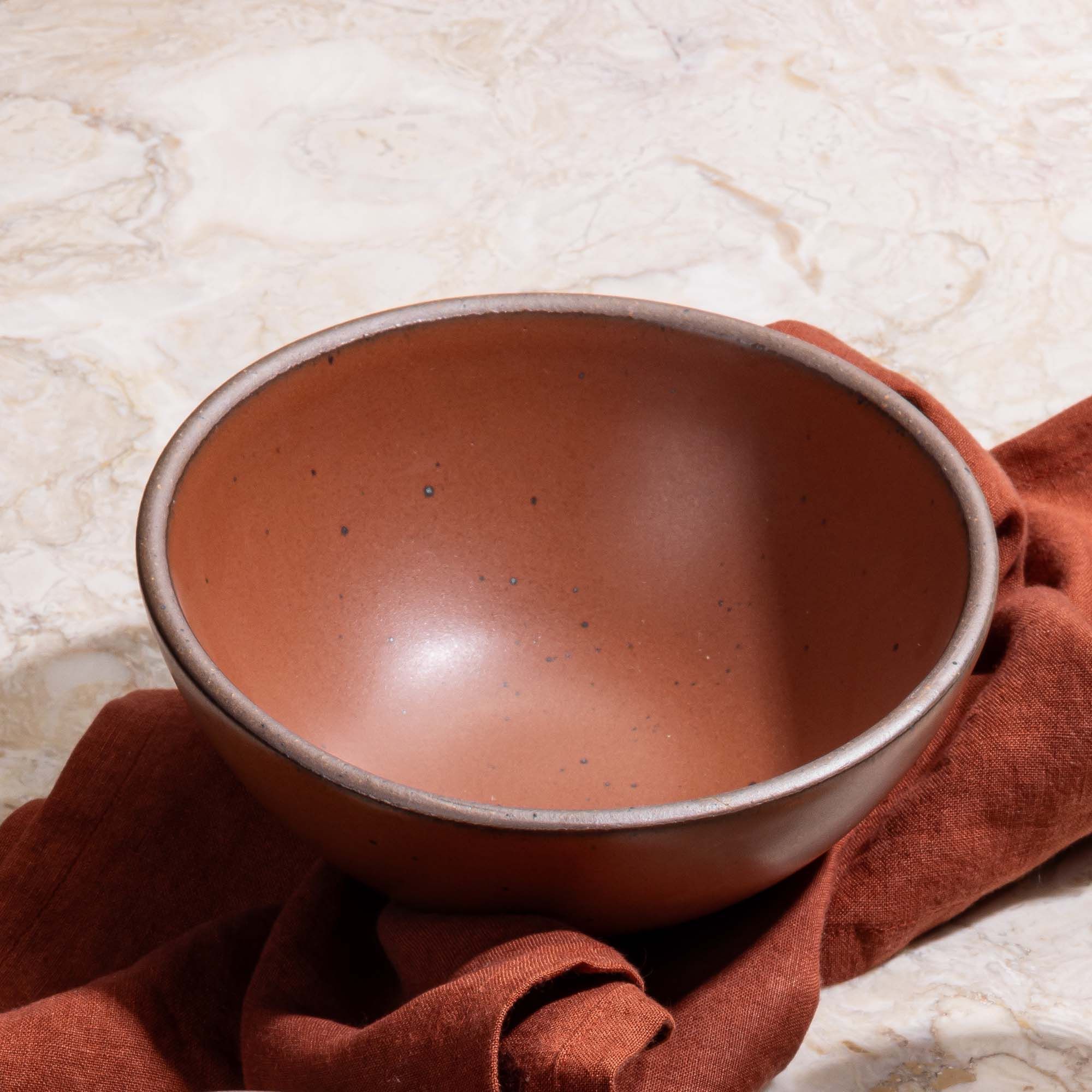 A medium rounded ceramic bowl in a cool burnt terracotta color with a dinner napkin in the same color