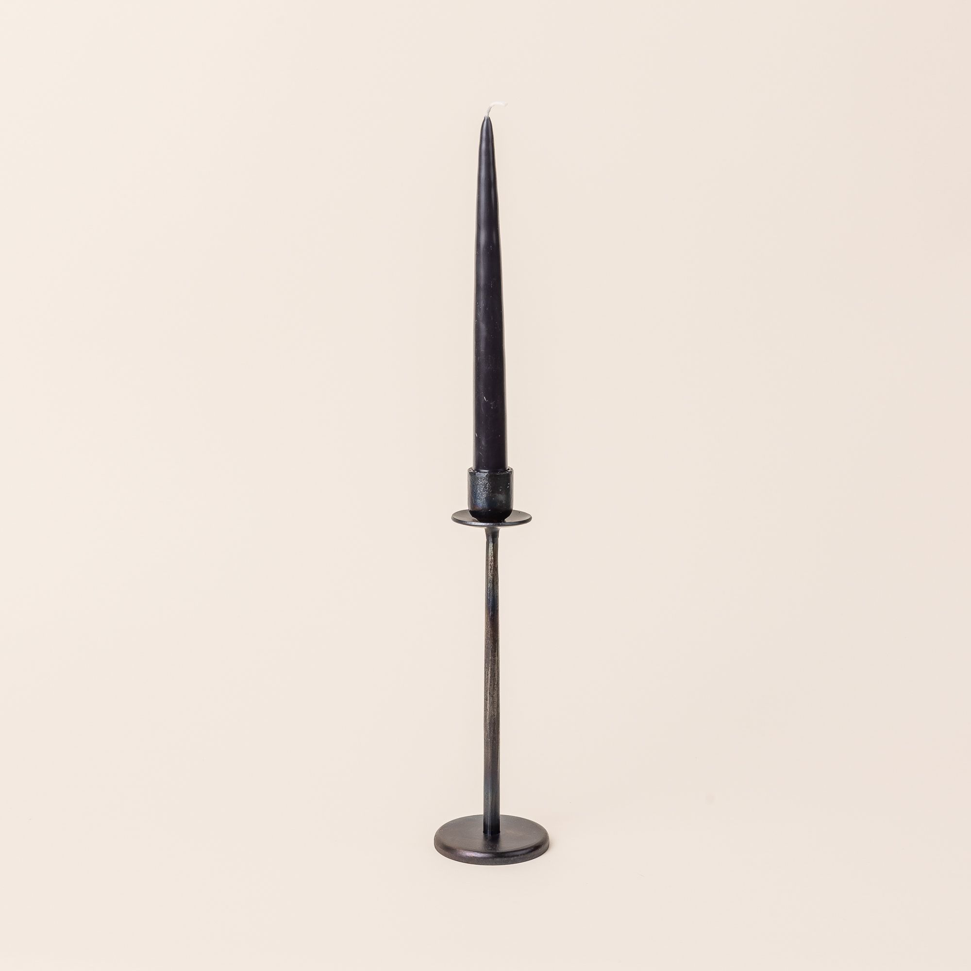A black taper candle sits on a tall narrow iron candlestick holder. The holder has with a flat circle base and topped with a vessel to hold a taper candle.