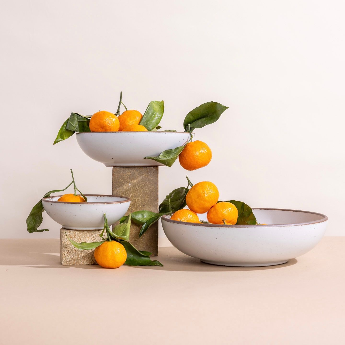 Three ceramic bowls in a cool white color holding oranges with green leaves, arranged on stone blocks against a light background