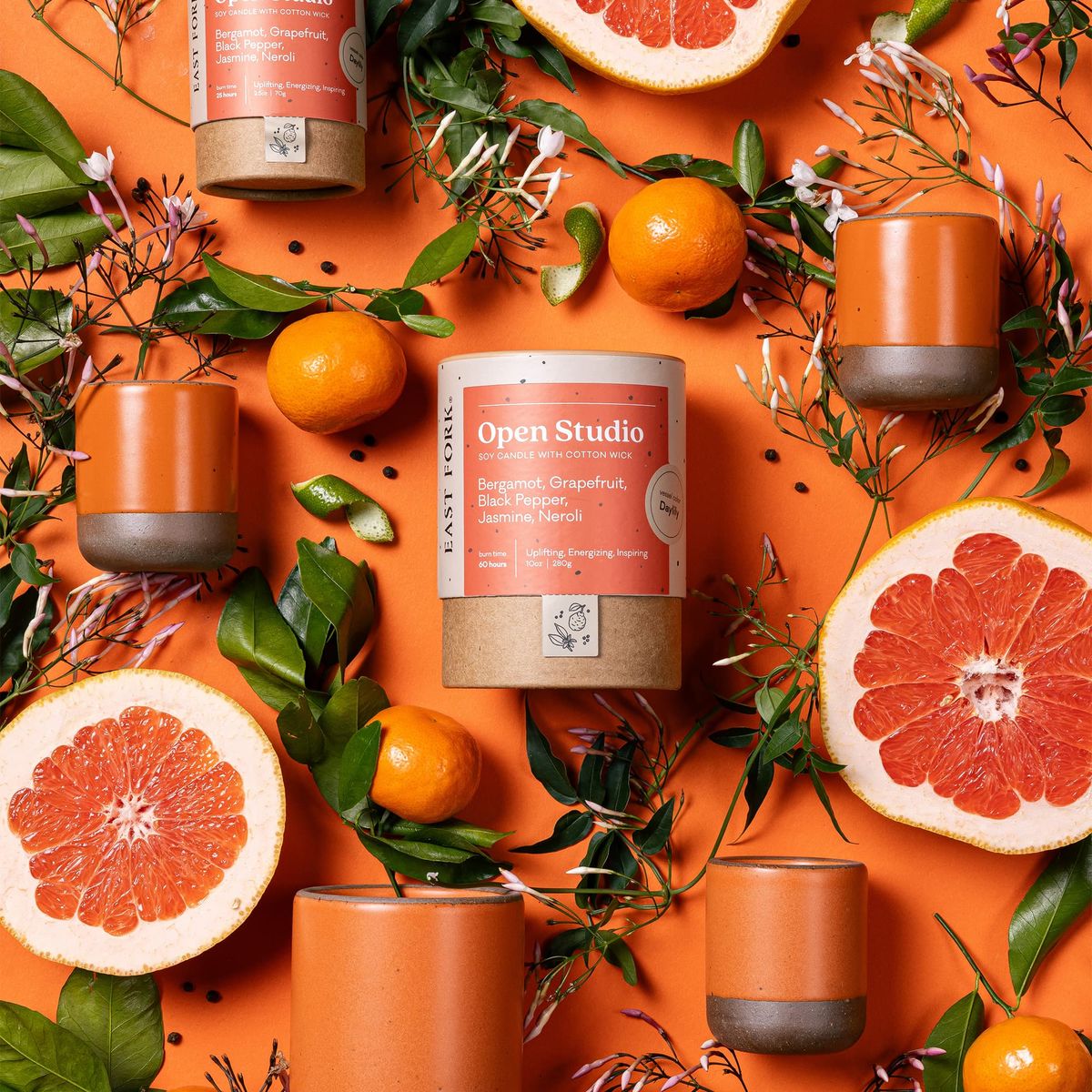 An artful layout of The Candle in Open Studio - centered is a cardboard packaging tube with the candle's name, surrounded by small and large ceramic candles in a cylindrical vessel in a bold orange color, surrounded by sliced grapefruits, oranges, and greenery.
