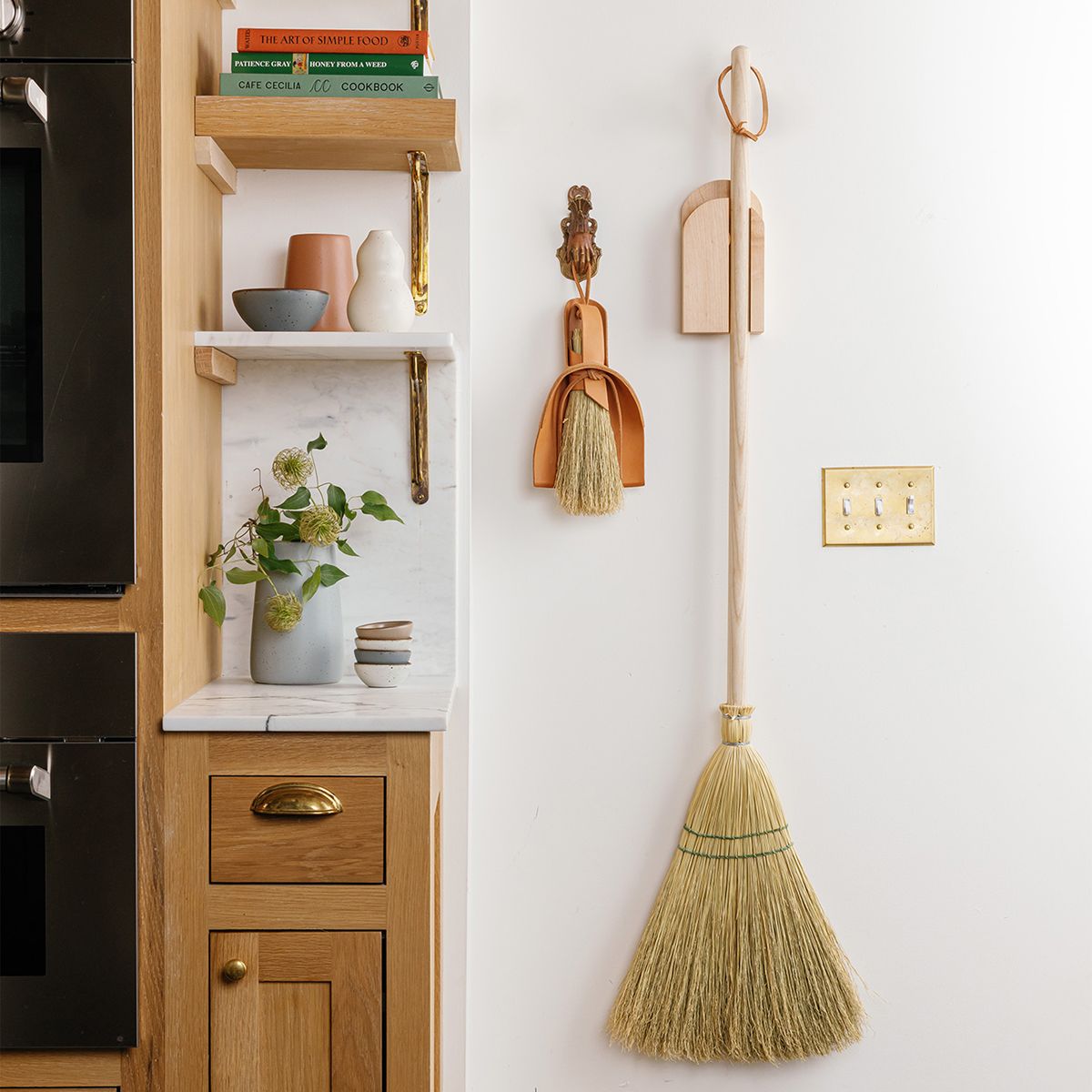 A rustic straw broom and smaller hand broom hang on a wall next to a styled shelf with vases and plant.