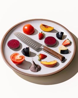 On a white ceramic plate, a santoku knife is artfully arranged in the center surrounded by colorful sliced fruits and vegetables.