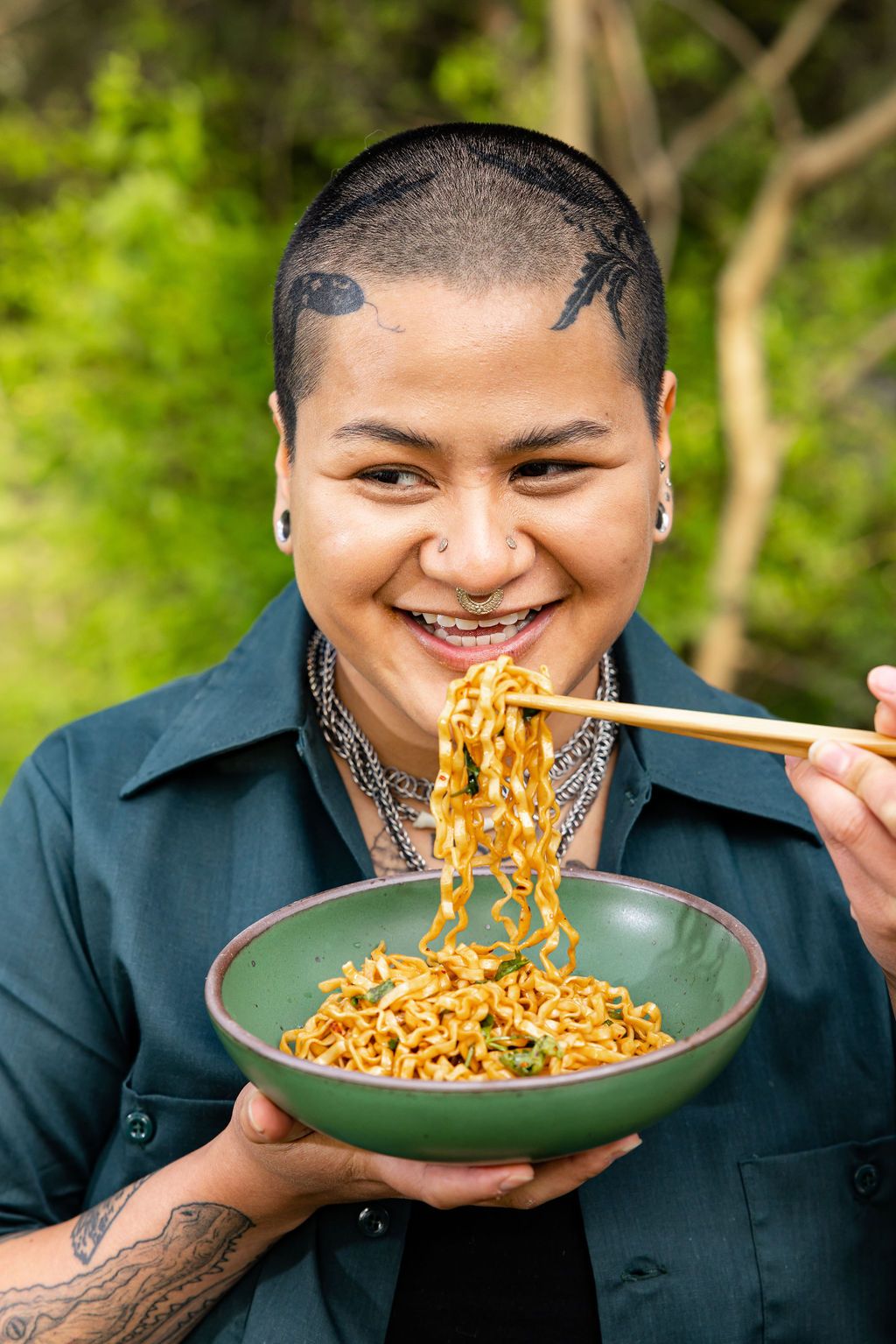 A person with buzzed hair and tattoos on his head and arm is smiling while holding noodles with chopsticks pulled from a medium green bowl.