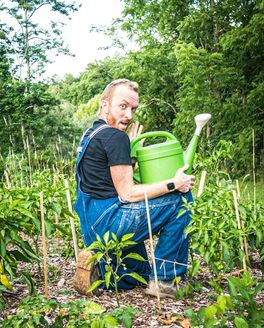 Outside, Donnie is kneeling on one knee wearing overalls and holding a watering can with a playful look