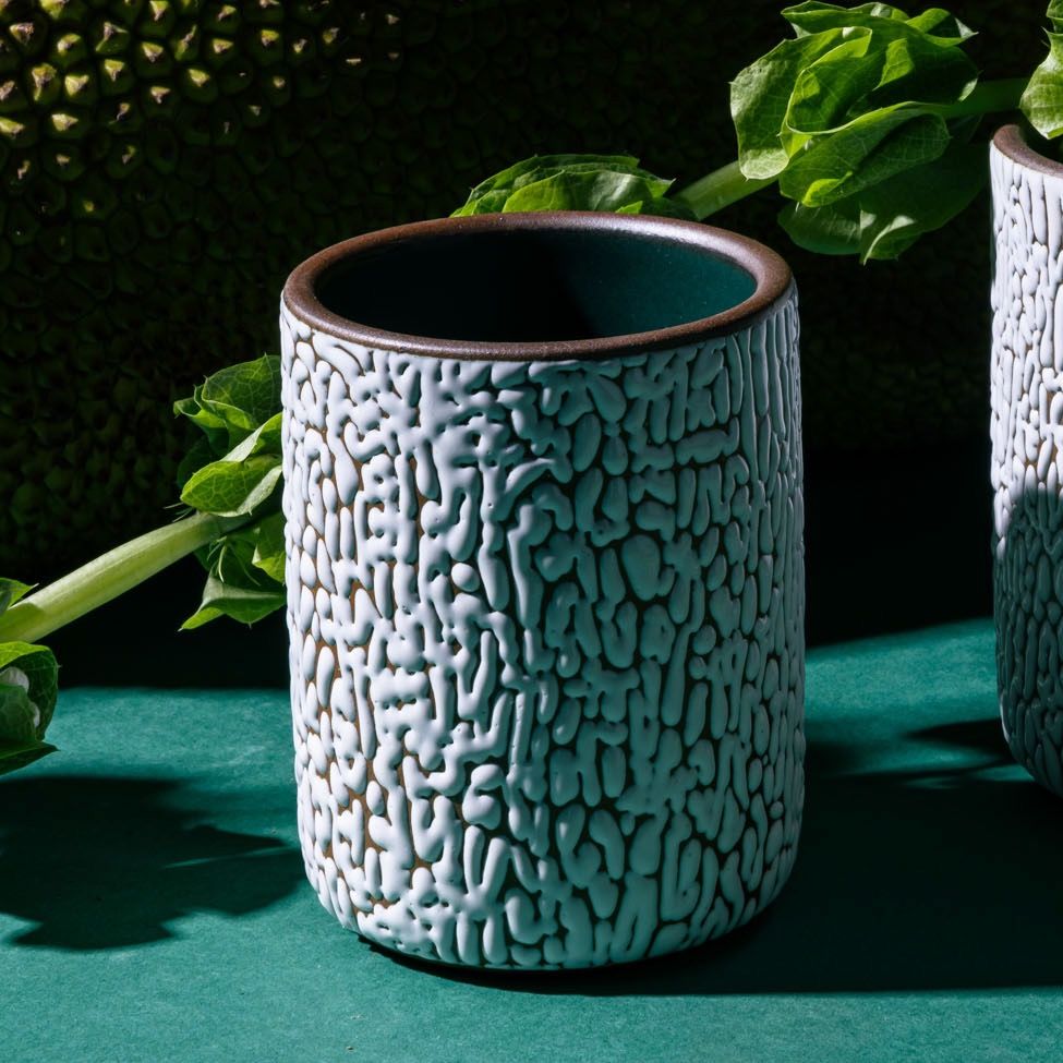 A medium white ceramic vessel with cracked texture and the interior being dark teal in a moody studio setting with foliage in the background