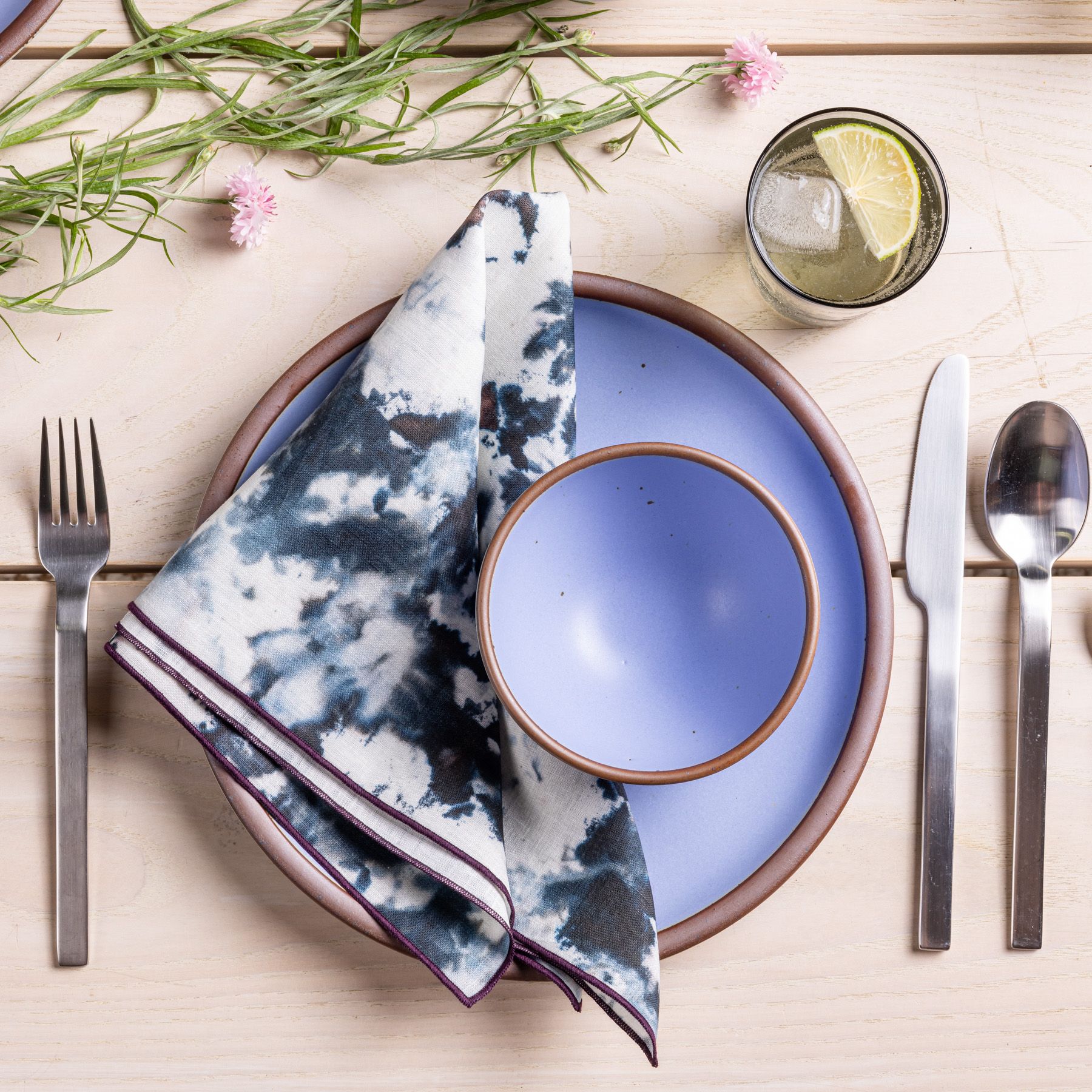 Top-down view of a place setting of periwinkle ceramic dinner plate and bowl with a triangle folded, marbled blue and white napkin. The setting is flanked with modern stainless steel flatware and a glass with water and a lemon wedge. All on a light cream colored wood table background.