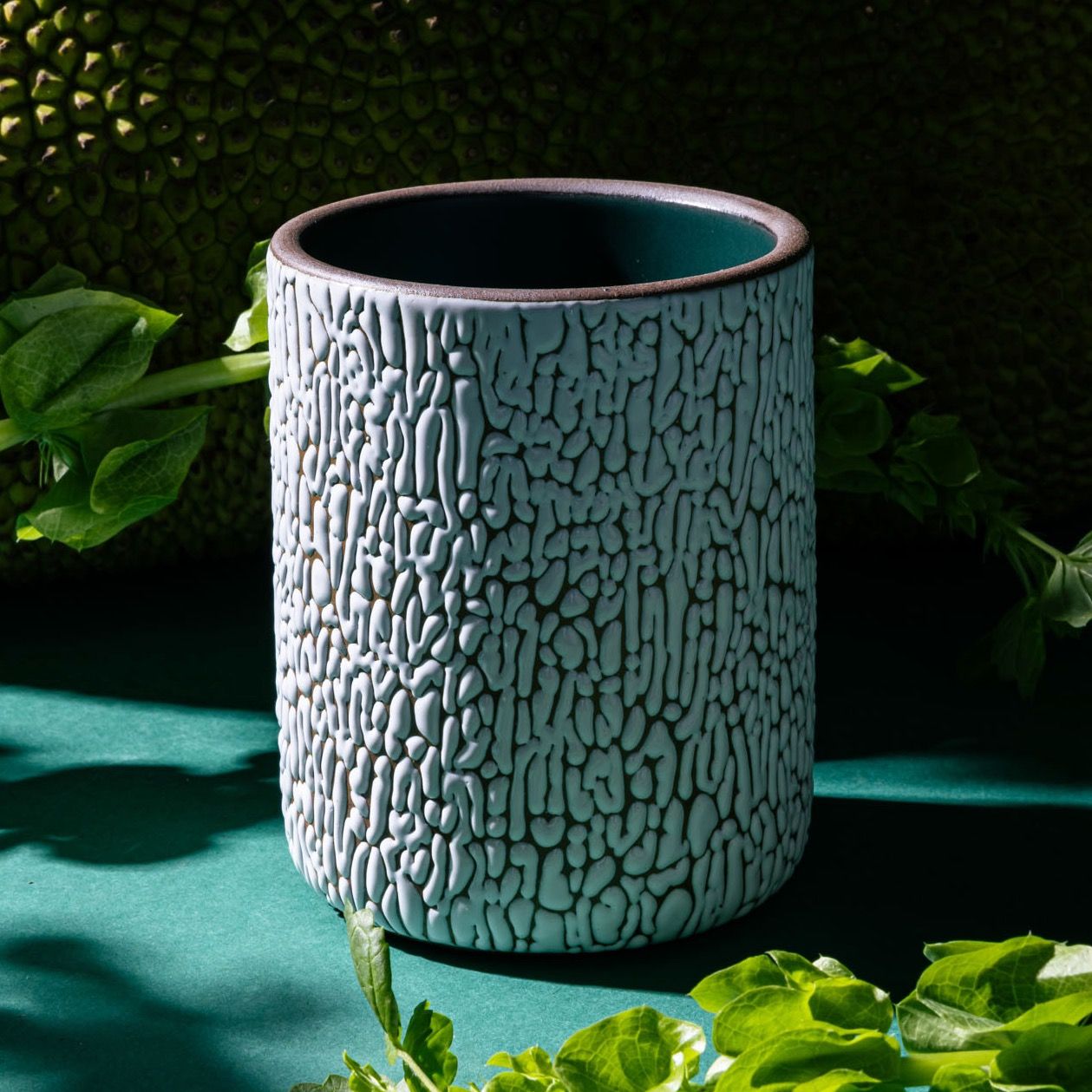 A big white ceramic vessel with cracked texture and the interior being dark teal in a moody studio setting with foliage in the background