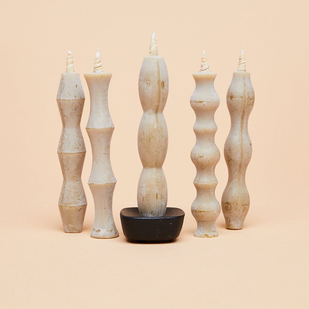 A group of five tan candles, taller than they are wide, each with a different pattern of flares and tapers, one standing on a black stand