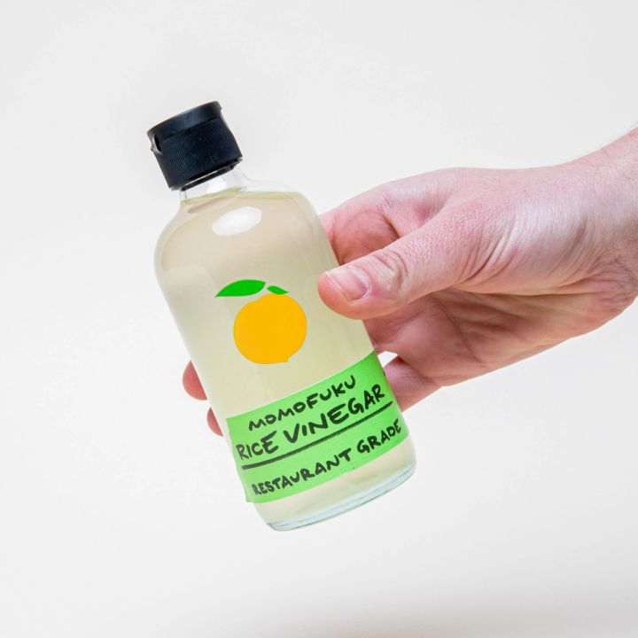 A hand holds a clear bottle with a black top filled with vinegar and a label that reads 'Momofuku Rice Vinegar"