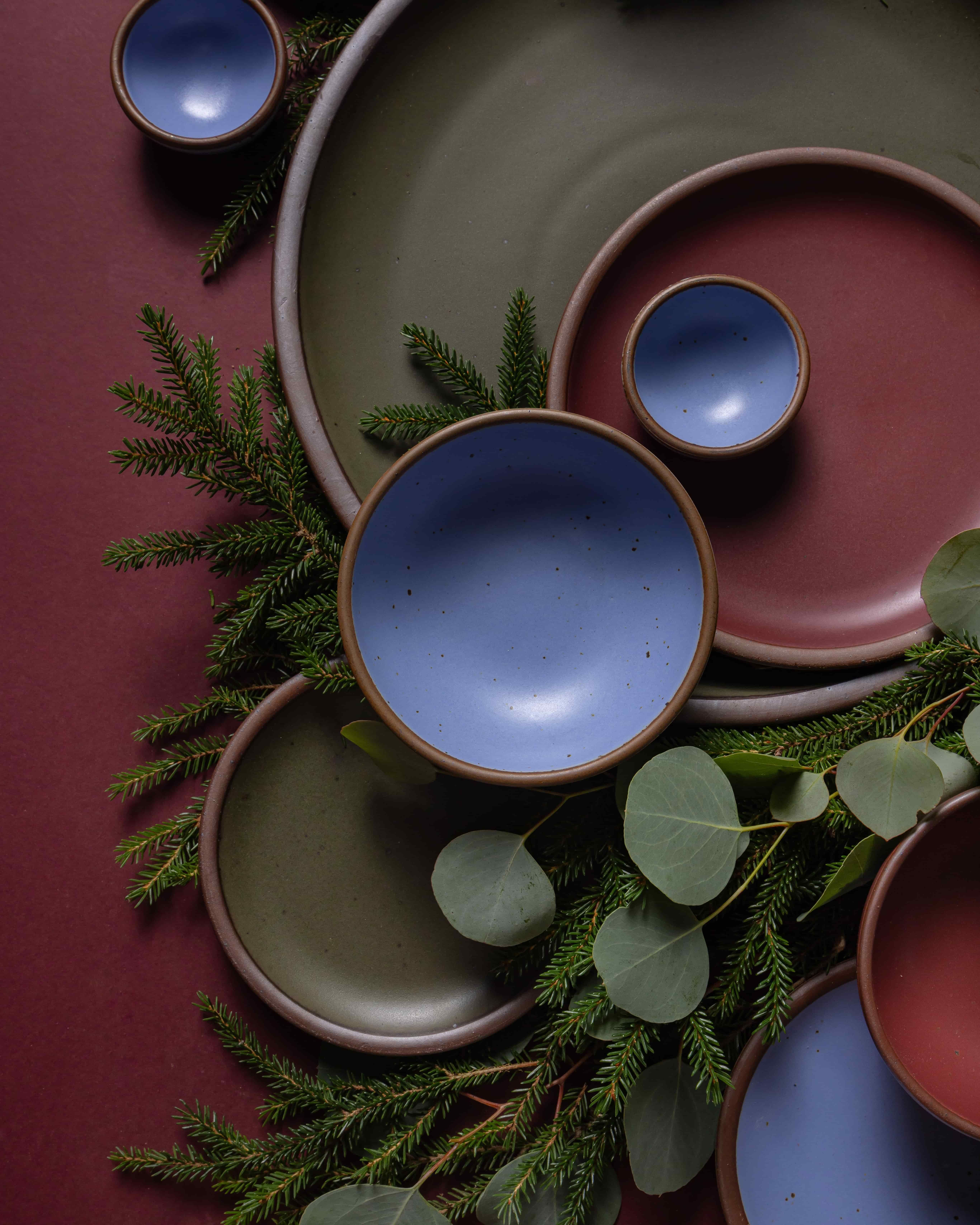 An artful arrangement of ceramic plates and bowls in forest green, periwinkle, and plum against a plum background and eucalyptus and pine leaves.