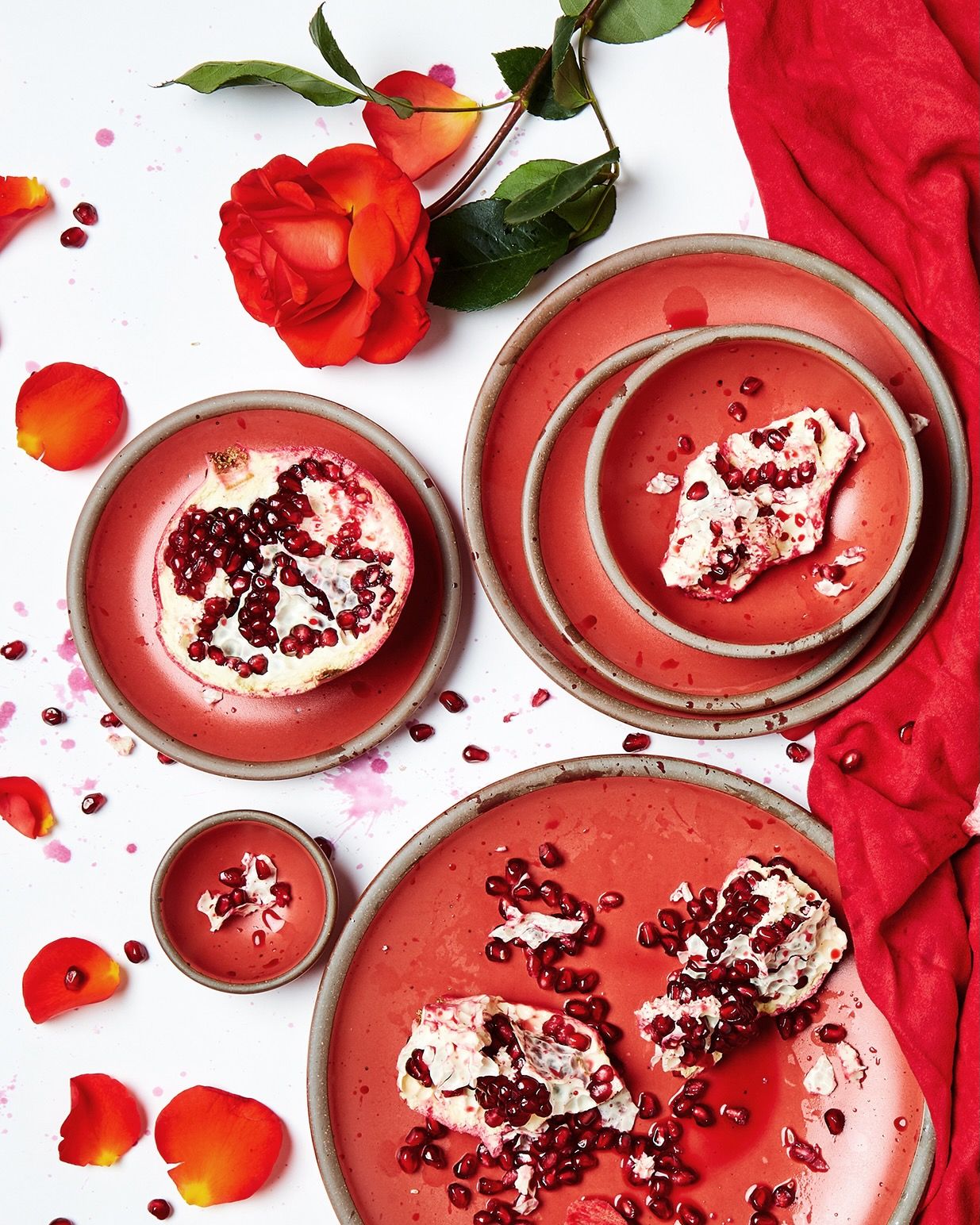 An overhead shot of ceramic plates in a bold red, artfully arranged with roses and rose petals, and pomegranates and pomegranate seeds.