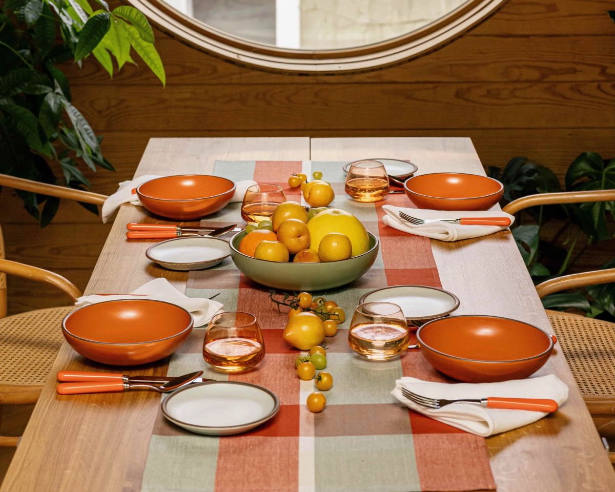 A table set with a gingham table runner, bold orange bowls and cool white plates, orange-handled flatware, whiskey snifters, and a centerpiece of fruits and vegetables. Also pictured is a woven chairs at the table table, a large houseplant, and a large circular window.