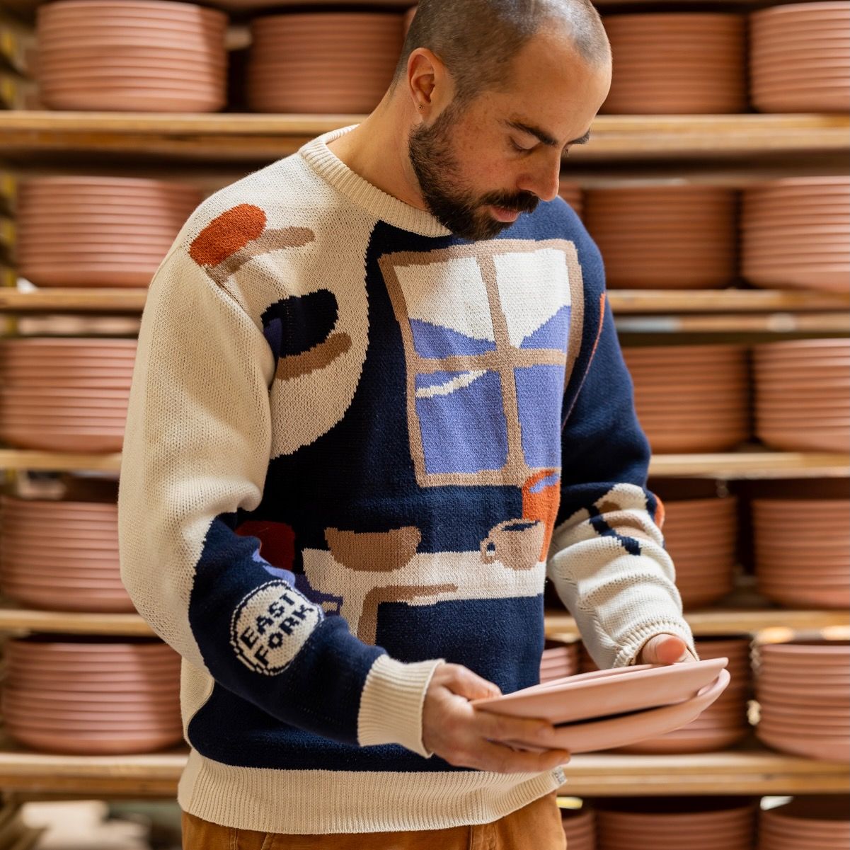 Alex wears a sweater with an illustration of a pottery workshop with a large window, shelves, and table with mugs and bowls. Alex holds 2 ceramic plates.