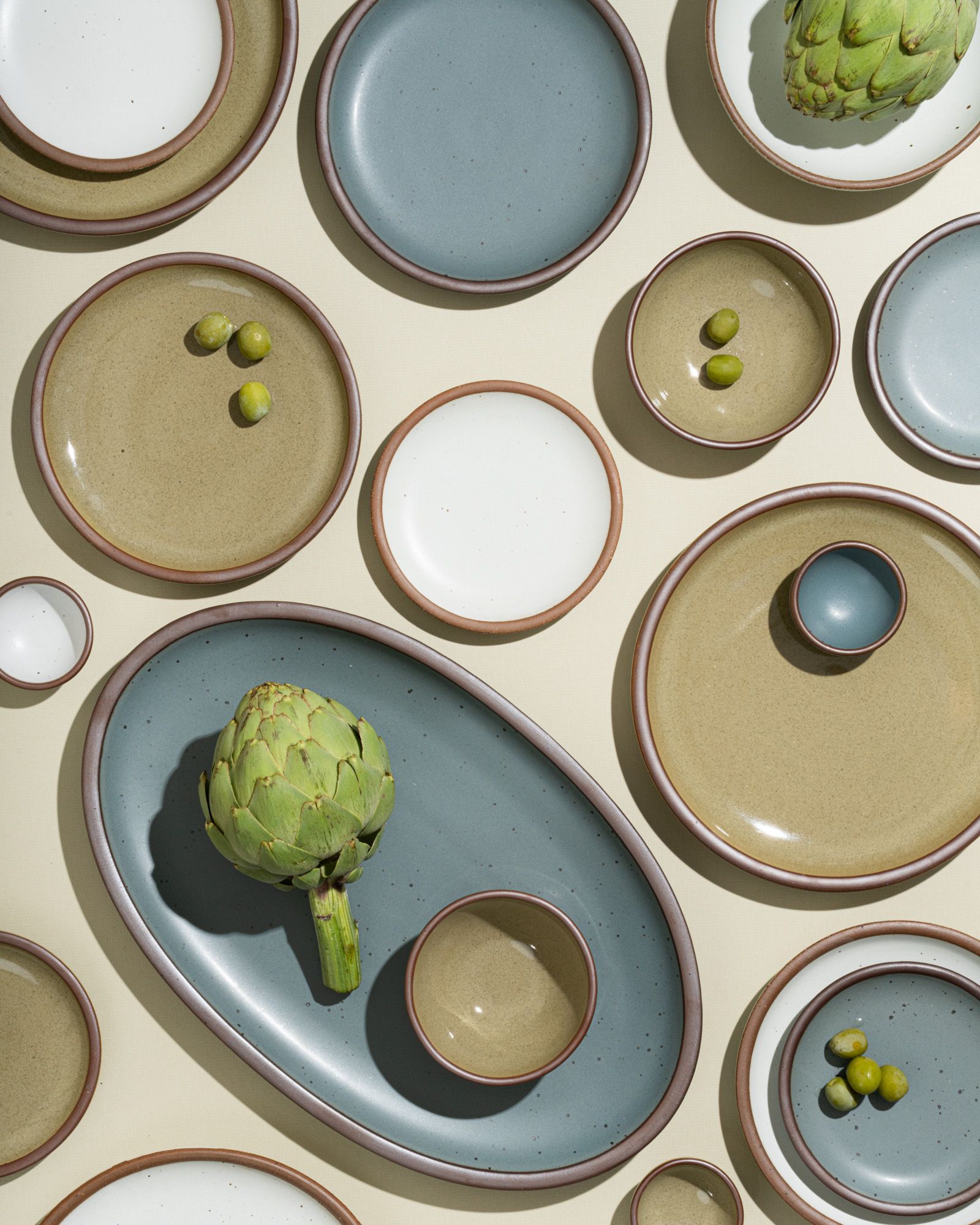 An overhead view of ceramic plates and shallow bowls in varying sizes in blue-grey, reflective dappled grey-green, and cool white styled with artichokes and olives.