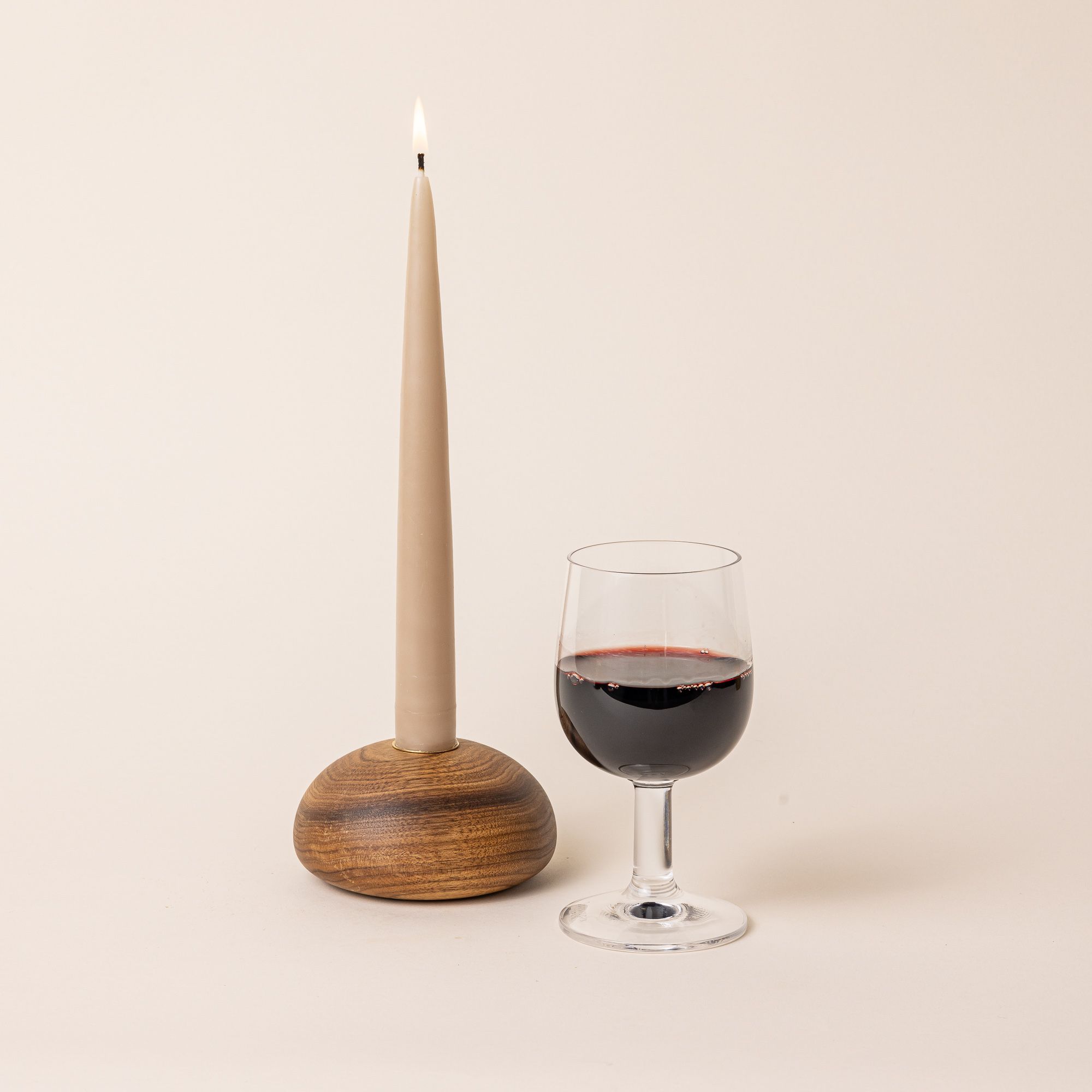 A clear wine glass with a short stem and wide cup, filled with red wine. Next to the glass is a lit tapered candle in a dome wood candlestick holder.