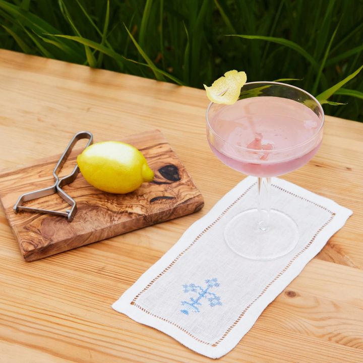 Outside on a wooden table is a pink cocktail in a martini coupe, sitting on a white cocktail napkin. To the left is a small cutting board with a peeled lemon and a stainless steel peeler.