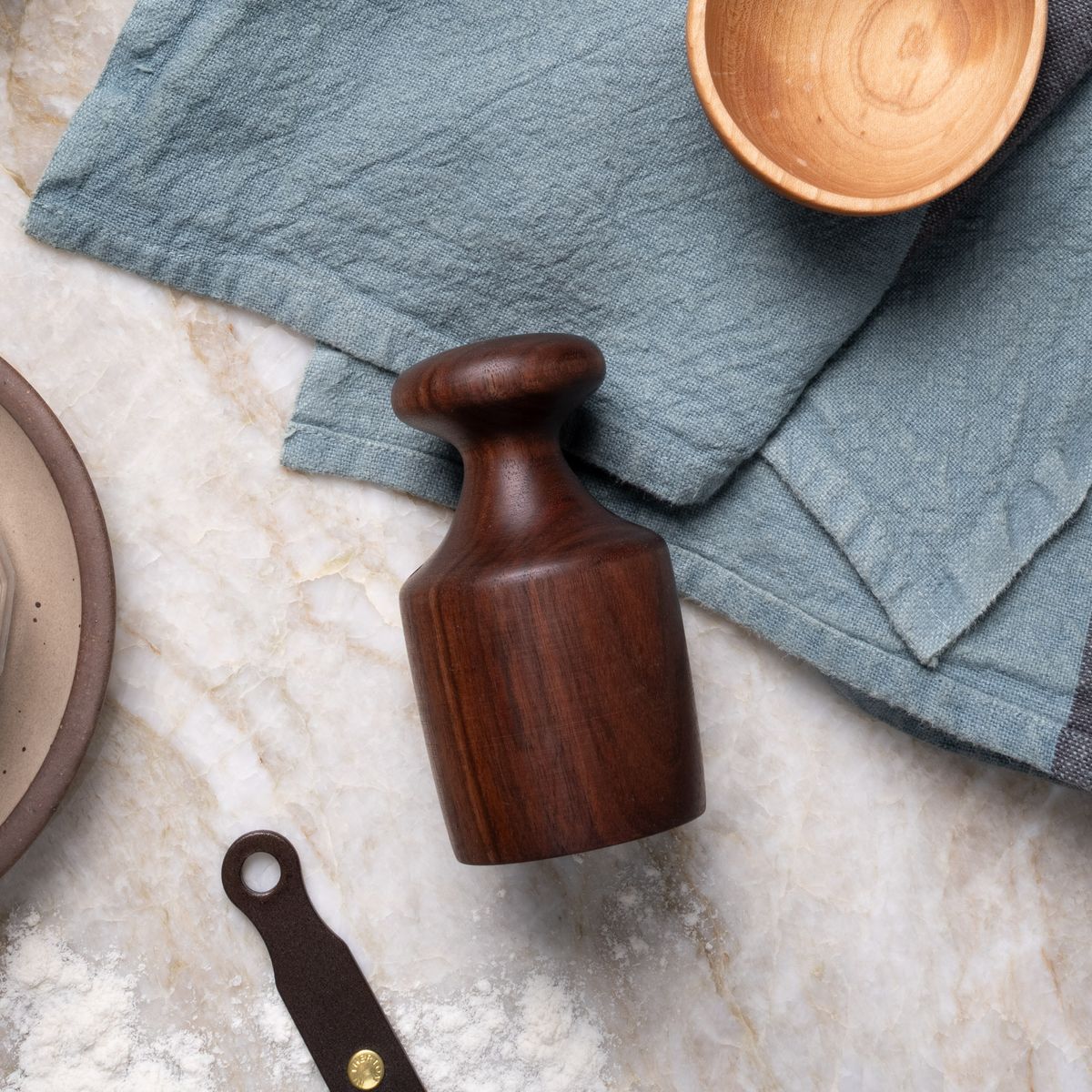 Dark wooden dough tamper on a marble countertop, surrounded by a blue linen cloth, small wooden bowl, and a dusting of flour.