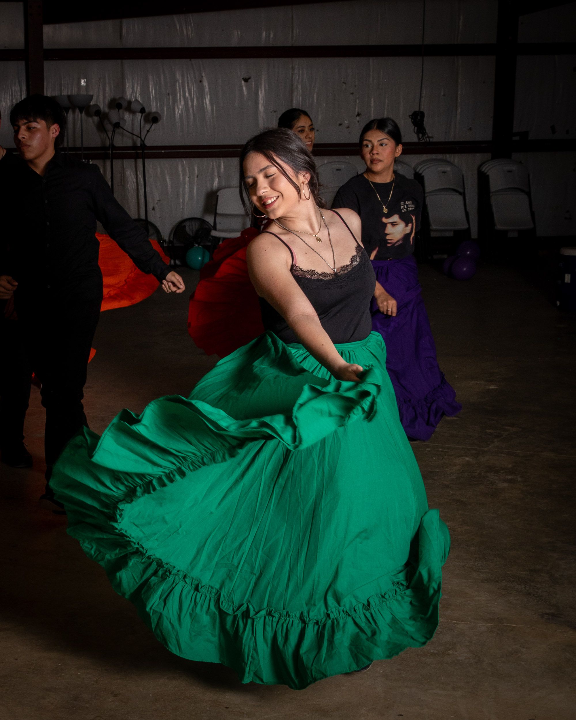 In a rehearsal space, a young woman is dancing with large colorful long skirt in green and smiling.