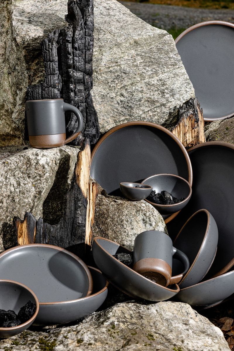 An group of grey plates, bowls, and mugs stacked haphazardly amongst large rocks and charred firewood. 