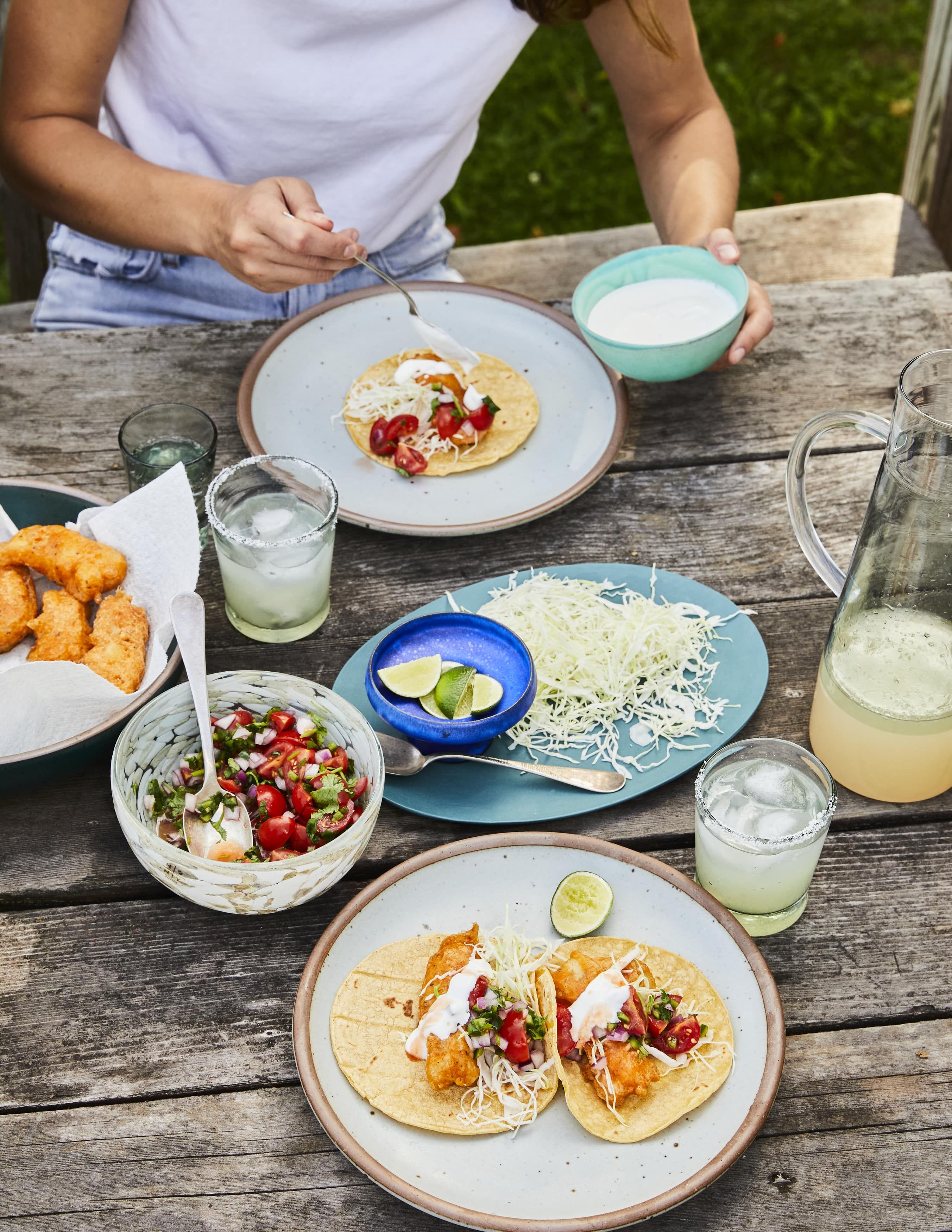 At an outdoor table, are fixings for baja fish tacos with a plate already ready with two tacos and a pitcher with glasses of margaritas