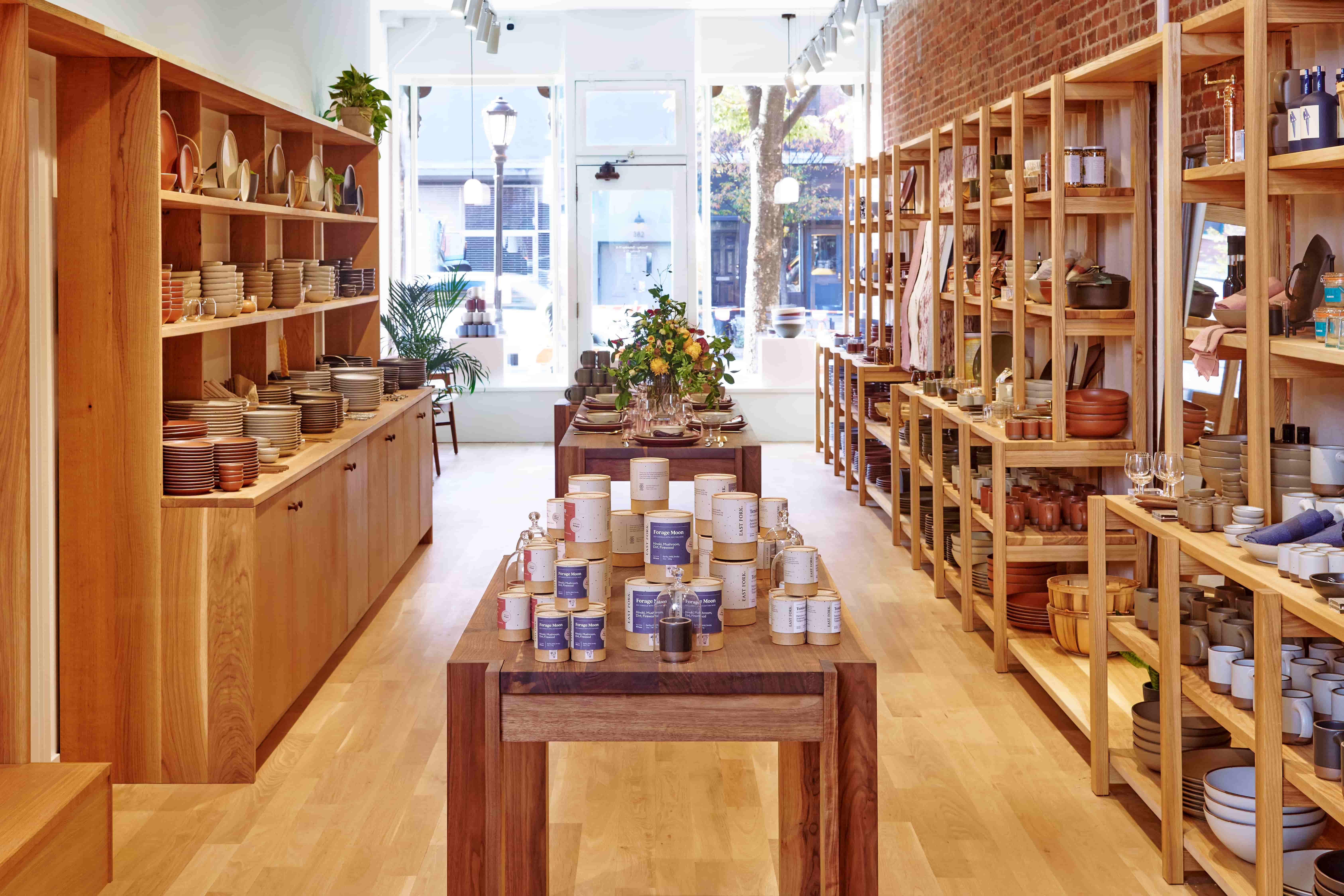 A view of East Fork Brooklyn from the back of the store with a long table down the middle and warm shelves filled with items like pottery, table linens, candles and more.