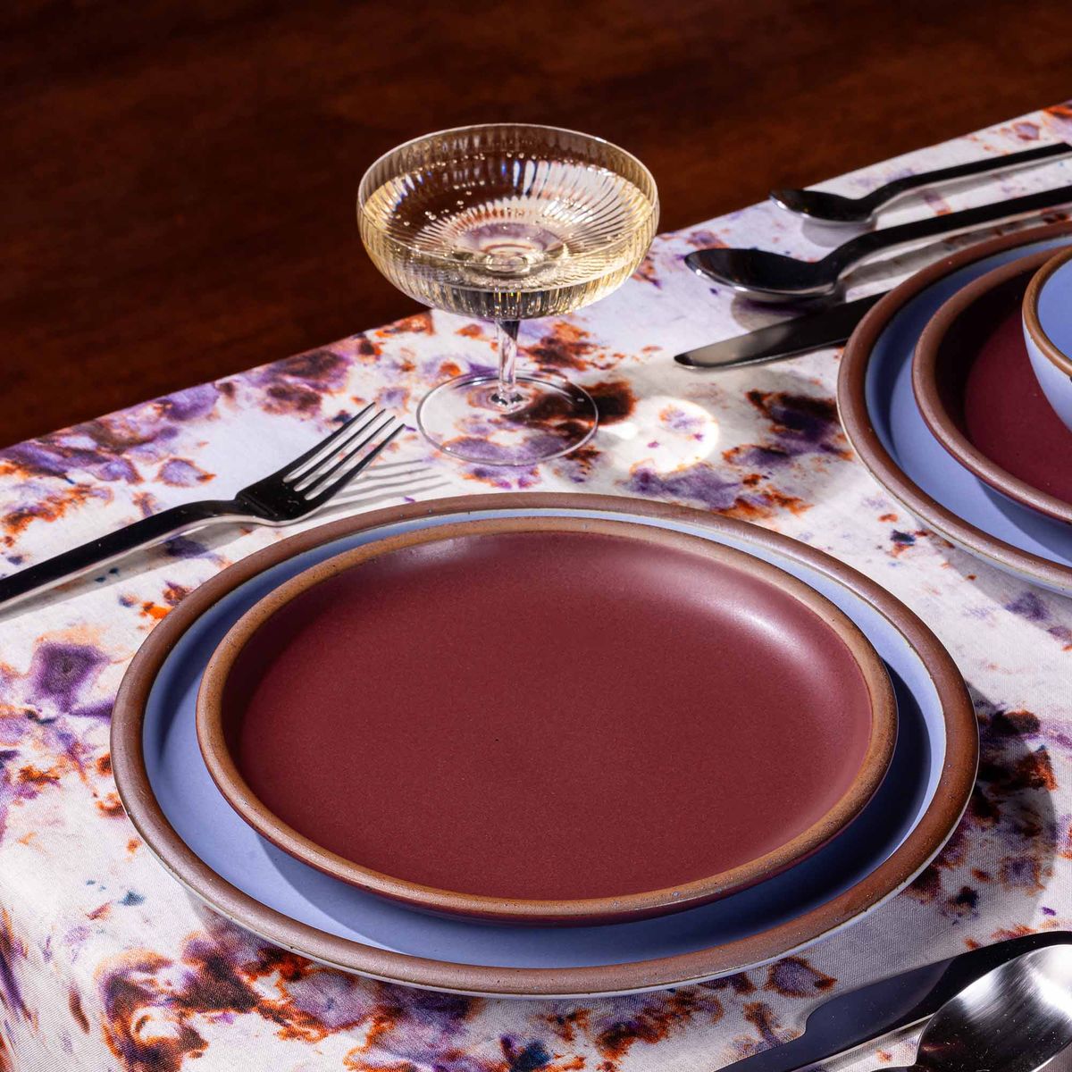 A table setting featuring marbled textiles, a coupe glass, and plates in plum and periwinkle colors