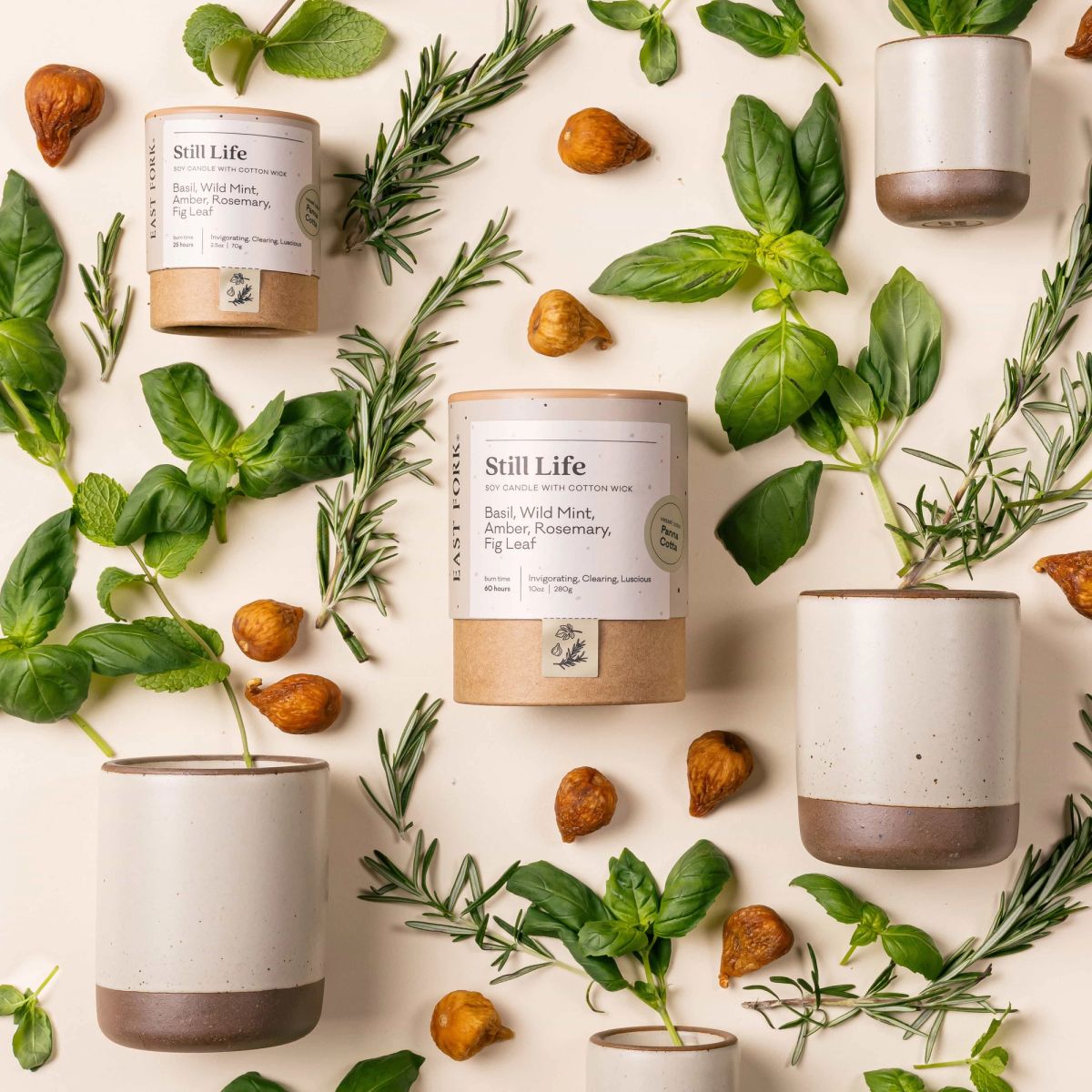 An artful layout of The Candle in Still Life - centered is a cardboard packaging tube with the candle's name, surrounded by small and large ceramic candles in a cylindrical vessel in a off-white color, surrounded by basil, rosemary, and fig leaf.
