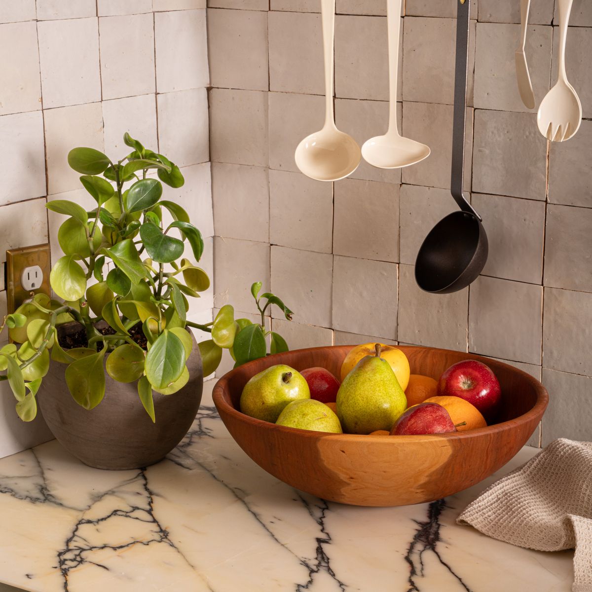 Cherry wood fruit bowl filled with pears, apples, and oranges on a marble countertop beside a leafy potted plant and hanging kitchen utensils.
