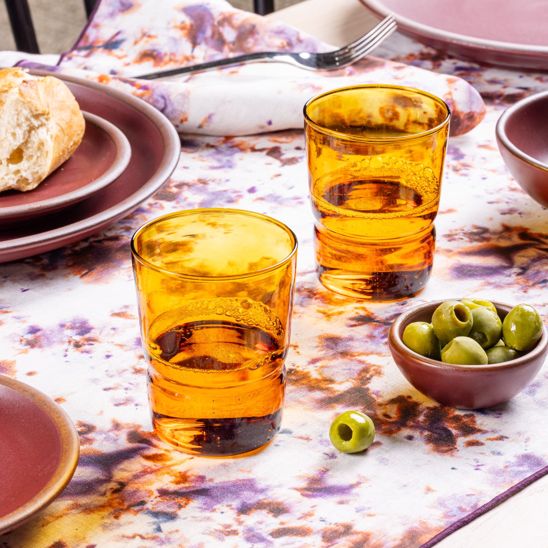 2 tumbler glasses that widen at the top in an amber color on a table with a plum marbled tablecloth and plum ceramic plates.