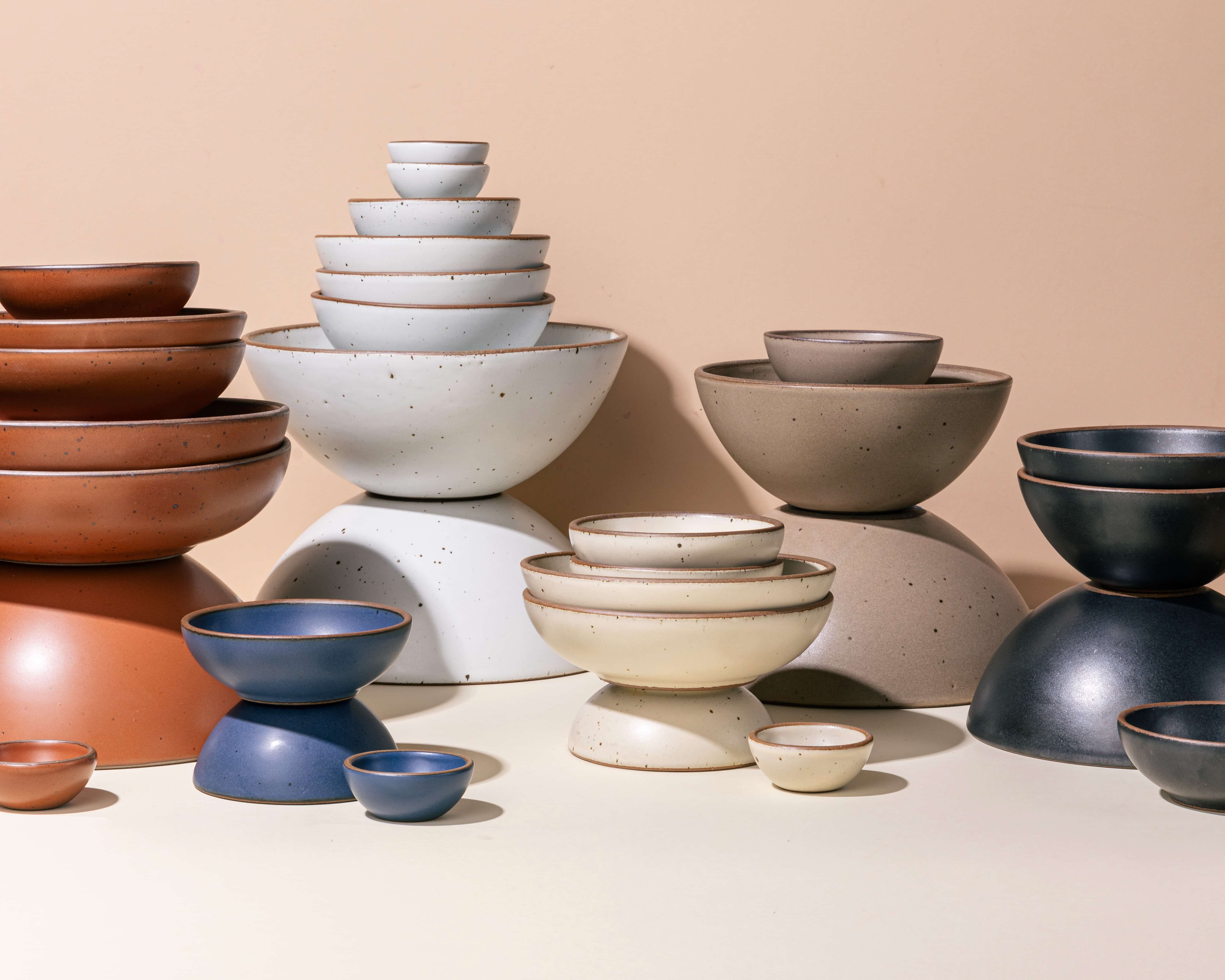 Bowls of various sizes and shapes are artfully arranged and stacked in colors including a cool terracotta, cool white, off-white cream, pale warm brown, cool medium blue, and graphite.