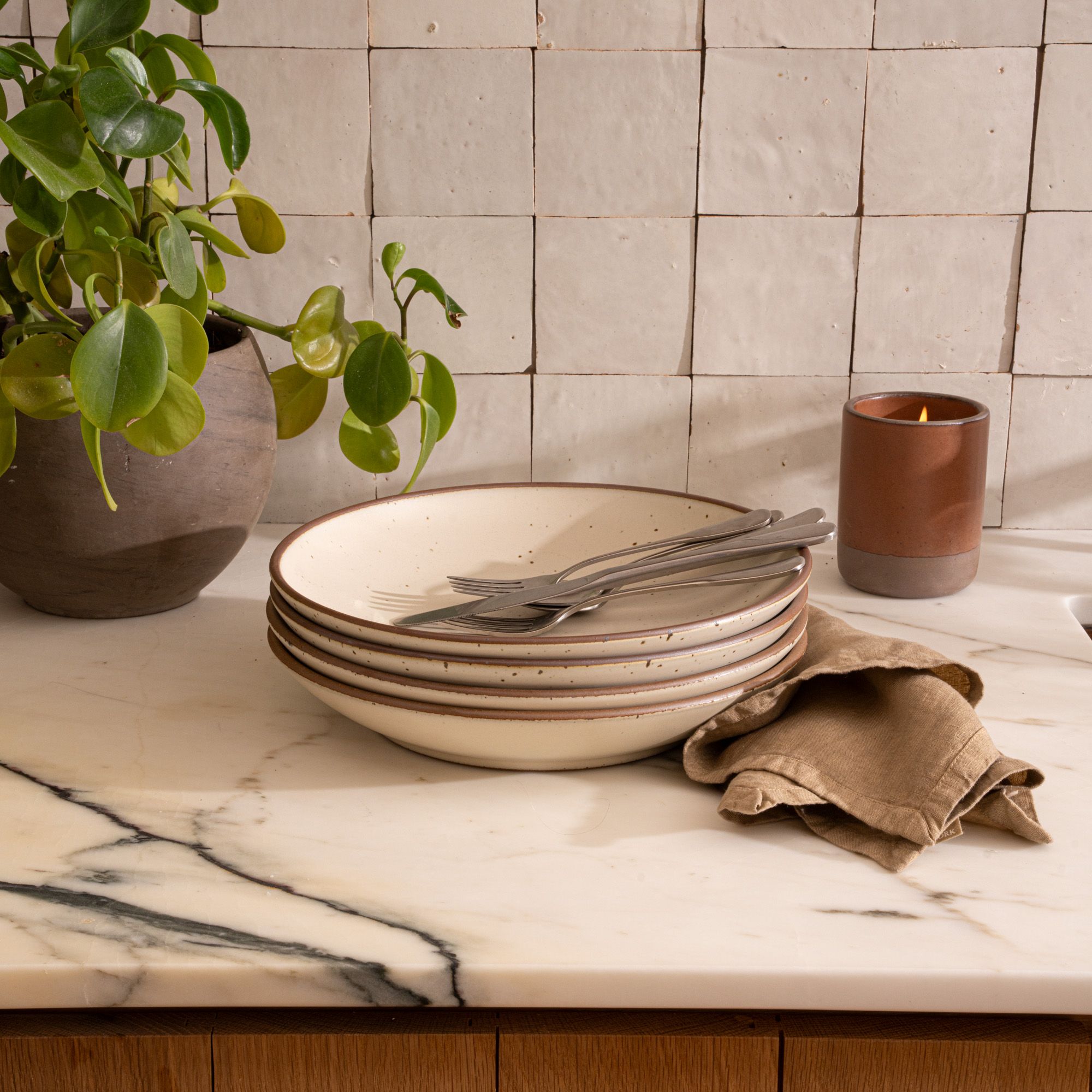 On a marble countertop is a stack of 4 large ceramic plates with a curved bowl edge in a warm, tan-toned, off-white color featuring iron speckles and an unglazed rim.